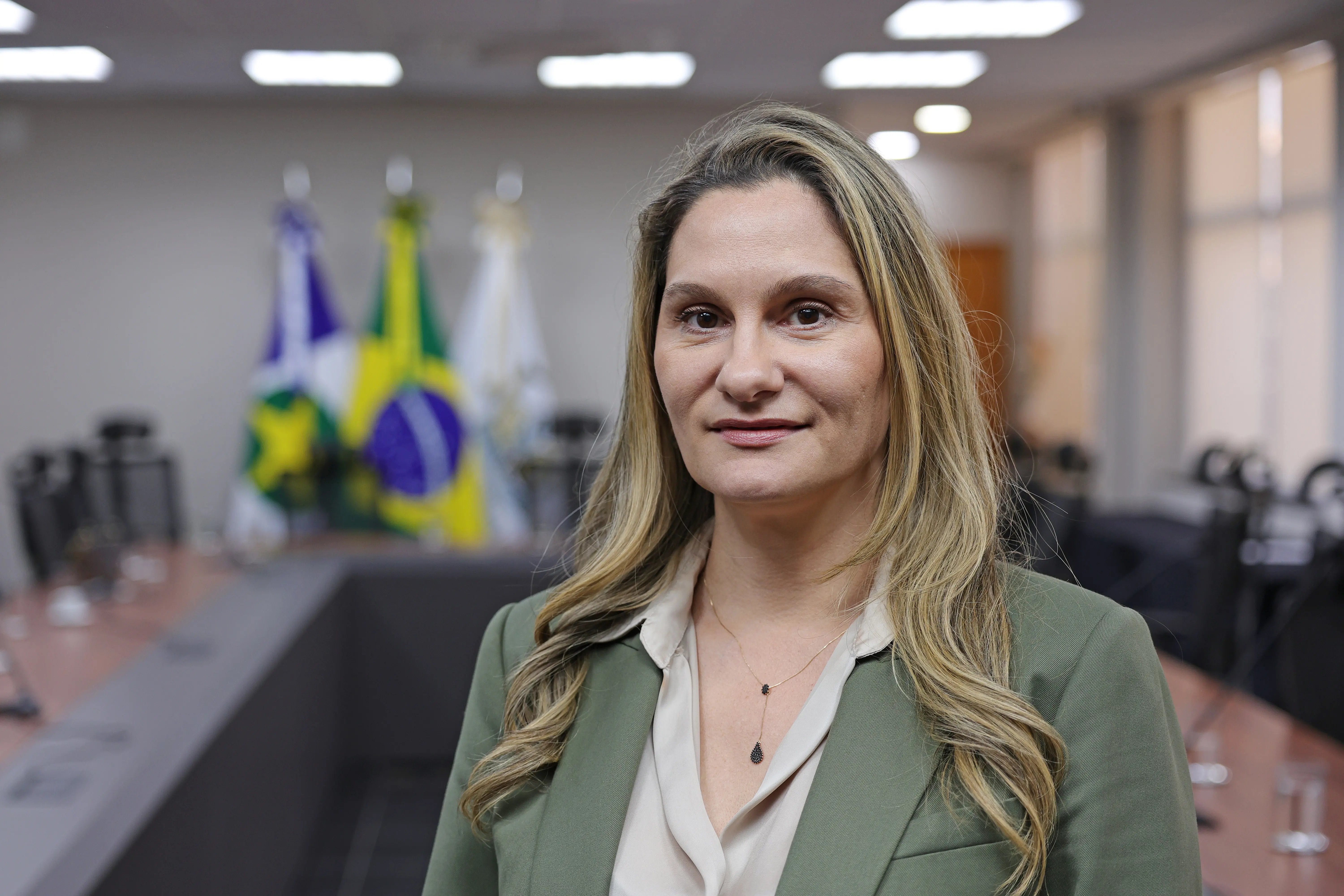 Retrato de uma mulher de cabelos loiros e longos trajando um blazer verde-oliva e blusa clara com colar fino. Ela olha para a câmera em um ambiente corporativo desfocado ao fundo.
