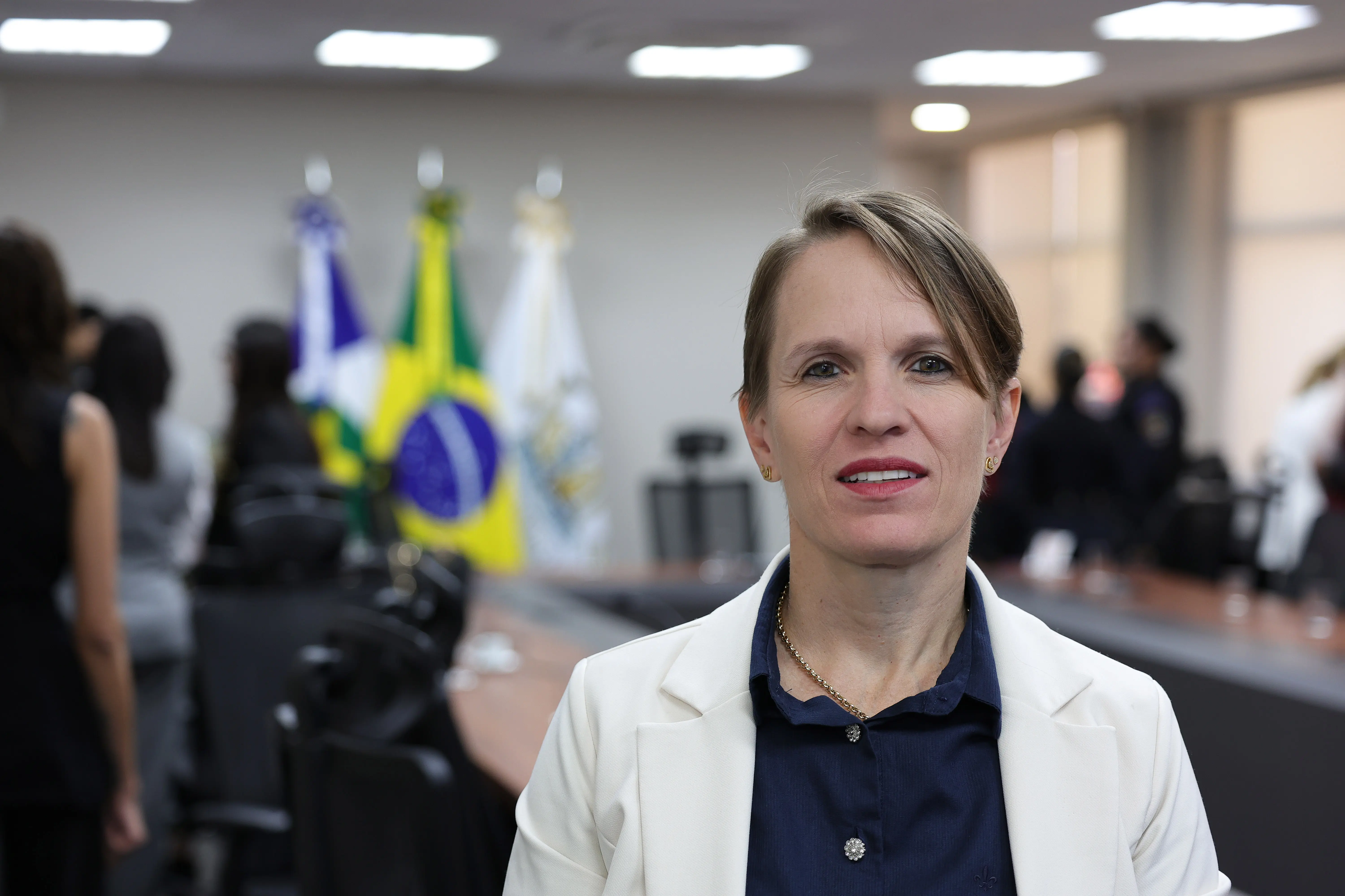 Retrato em plano médio de uma mulher de cabelo curto castanho claro, trajando blazer branco e camisa azul-marinho. Ao fundo desfocado, observam-se bandeiras oficiais e outras pessoas na sala.