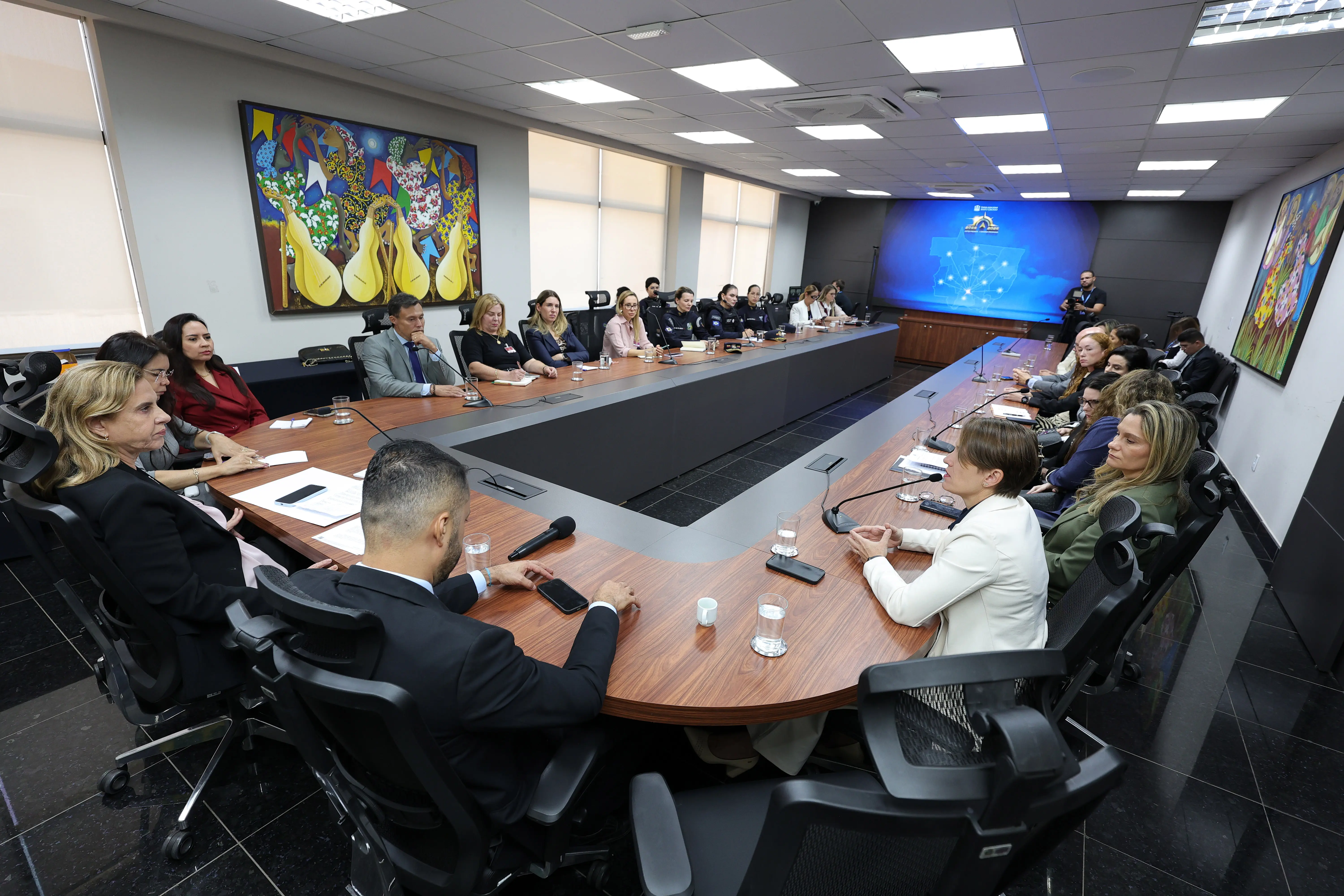 ista ampla de uma sala de reuniões com pessoas acomodadas em uma grande mesa retangular retada ao centro. Nas paredes há quadros coloridos e um painel digital ao fundo. Copos e microfones estão dispostos sobre a mesa.