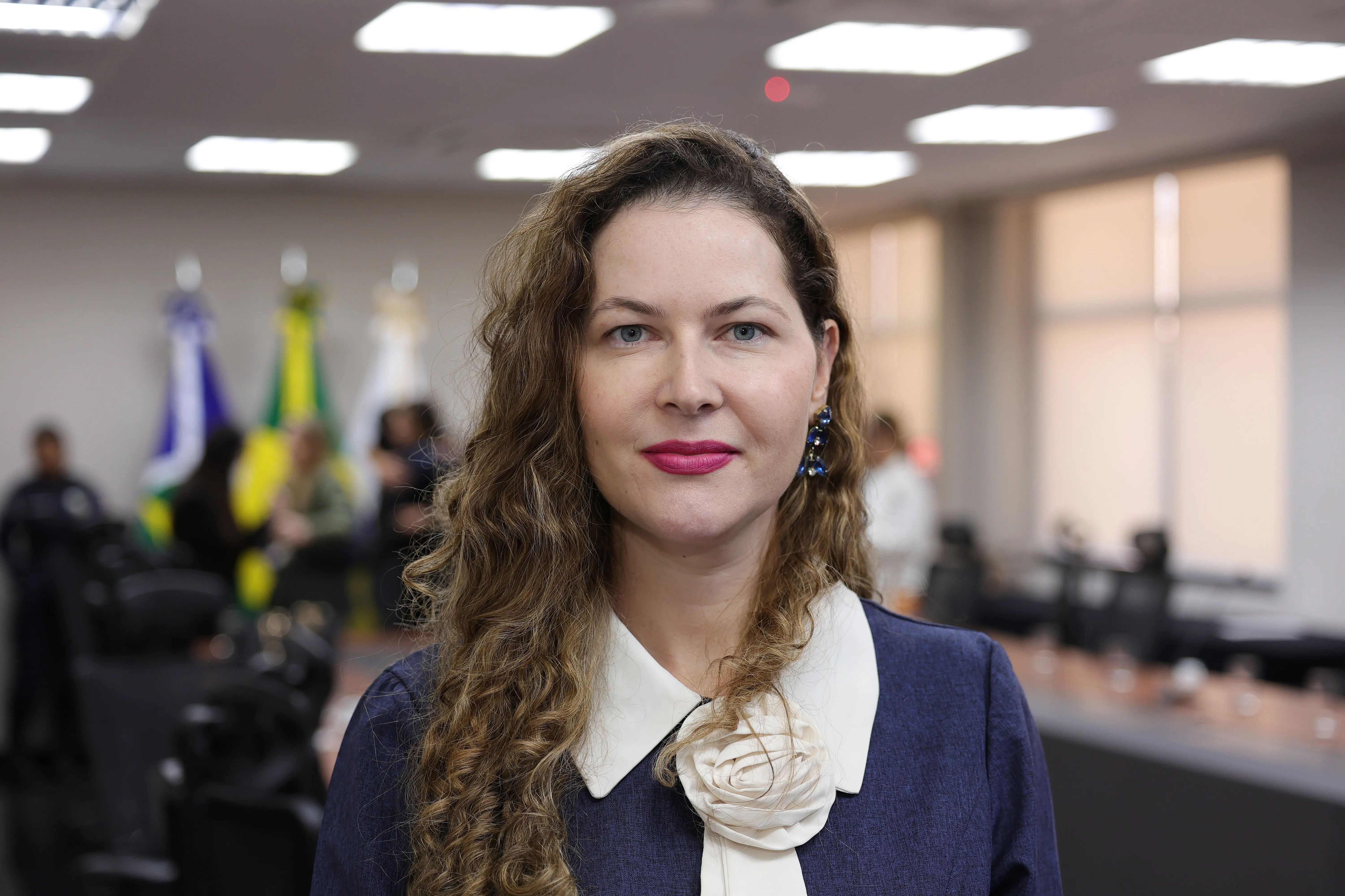 Retrato em plano médio de mulher de cabelos longos e ondulados, usando blazer azul-marinho sobre gola branca decorada com aplique de flor. O fundo desfocado mostra um ambiente corporativo com bandeiras.