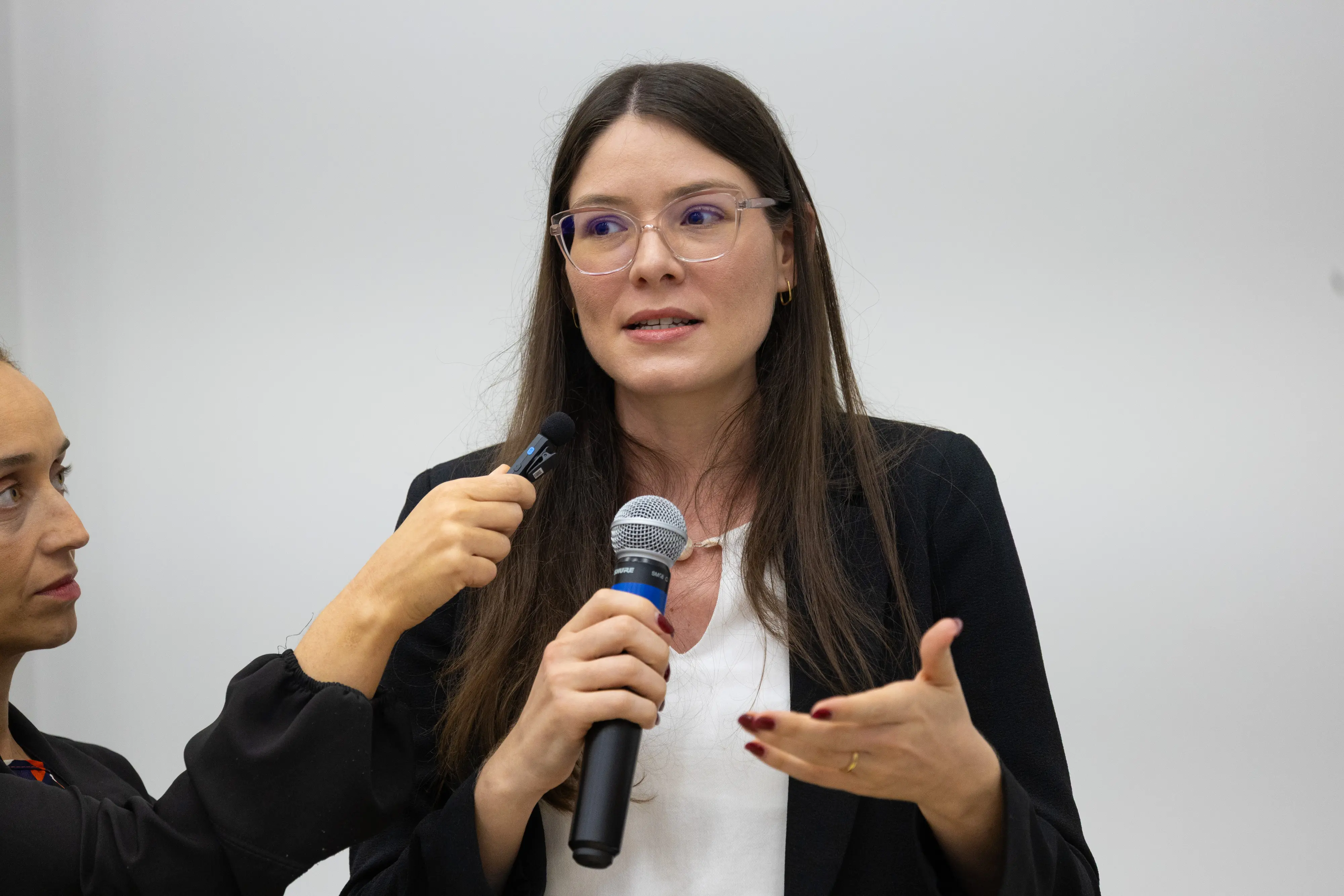 Mulher de óculos fala ao microfone durante oficina participativa. Pessoa ao lado segura gravador próximo à entrevistada.