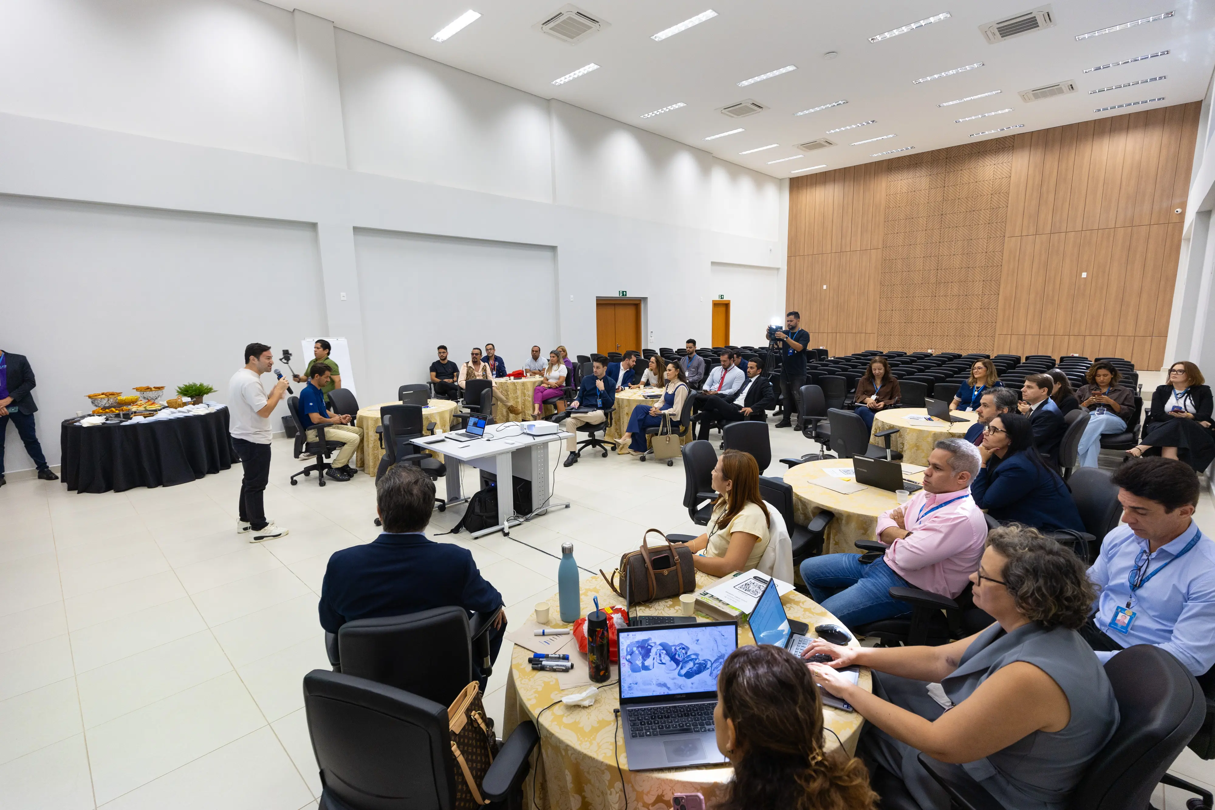 Participantes acompanham oficina em salão com mesas redondas e painel de madeira ao fundo.