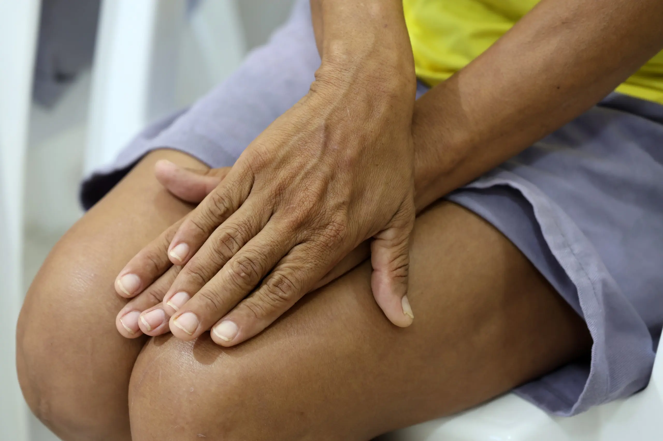 Close das mãos de uma mulher apoiadas sobre o joelho. Ela veste camiseta amarela e short cinza. A imagem destaca sinais de envelhecimento da pele e posição de espera.