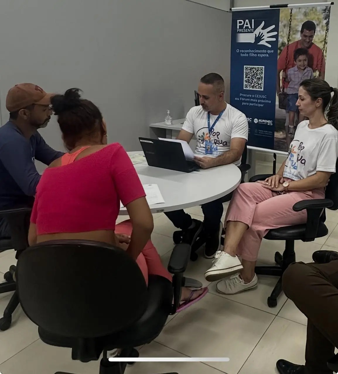 Quatro pessoas sentadas em volta de uma mesa redonda branca. Dois profissionais com camisetas 