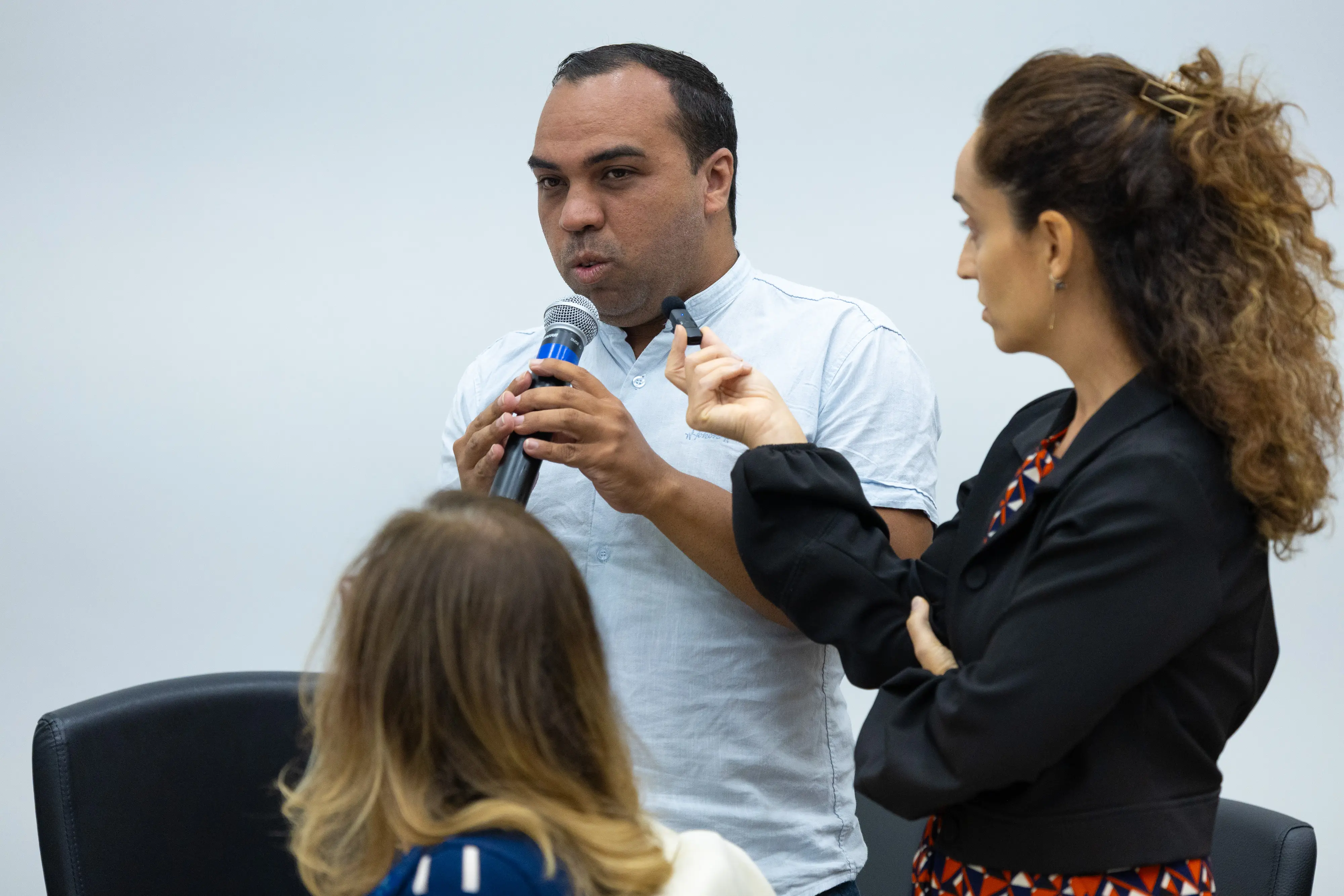 Homem de camisa clara fala ao microfone durante atividade participativa. Pessoas observam ao redor da mesa.