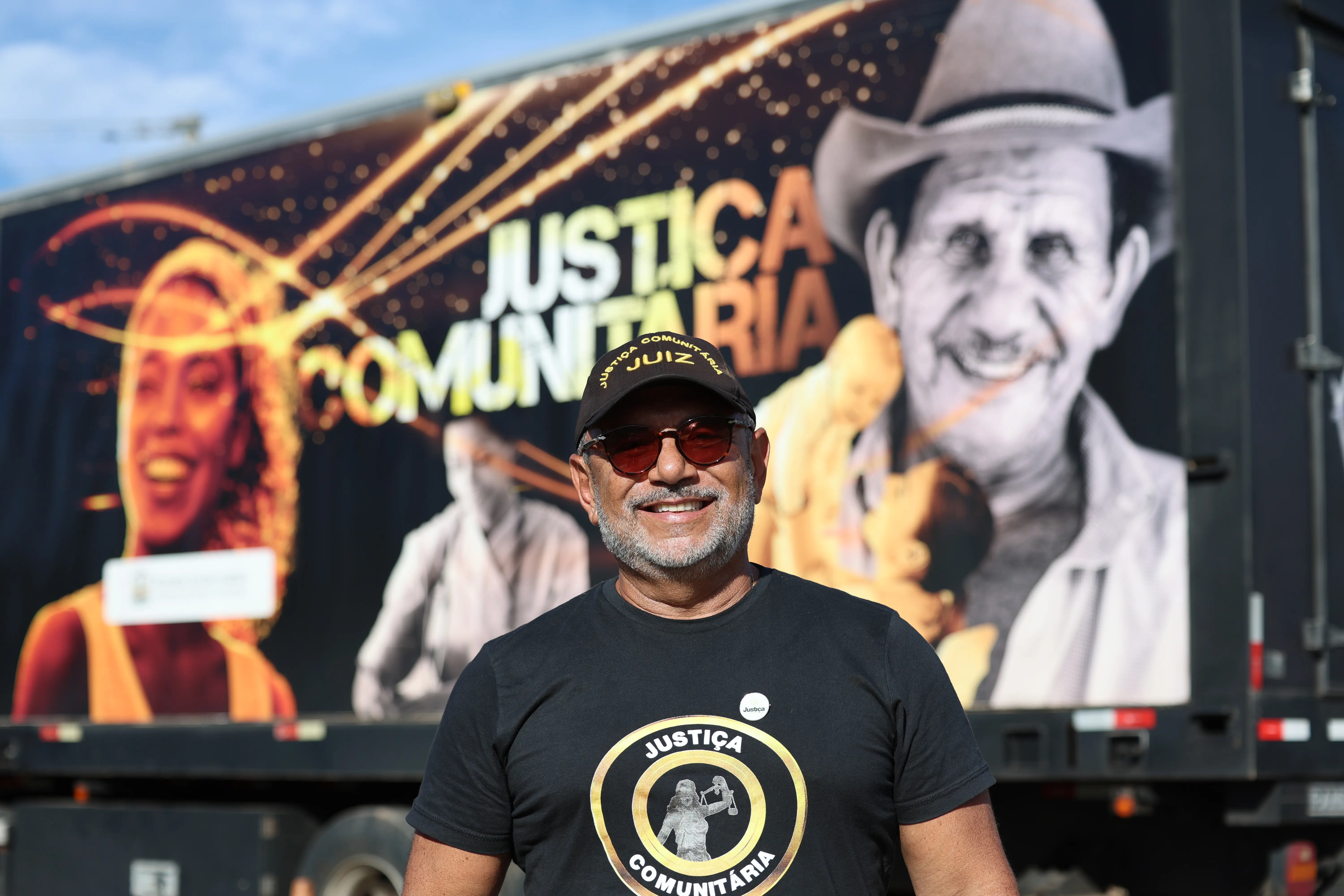 Foto do juiz José Antonio Bezerra Filho sorridente de camiseta e boné pretos escrito "Justiça Comunitária". Atrás dele, a lateral de um caminhão exibe uma arte colorida com o texto "Justiça Comunitária".
