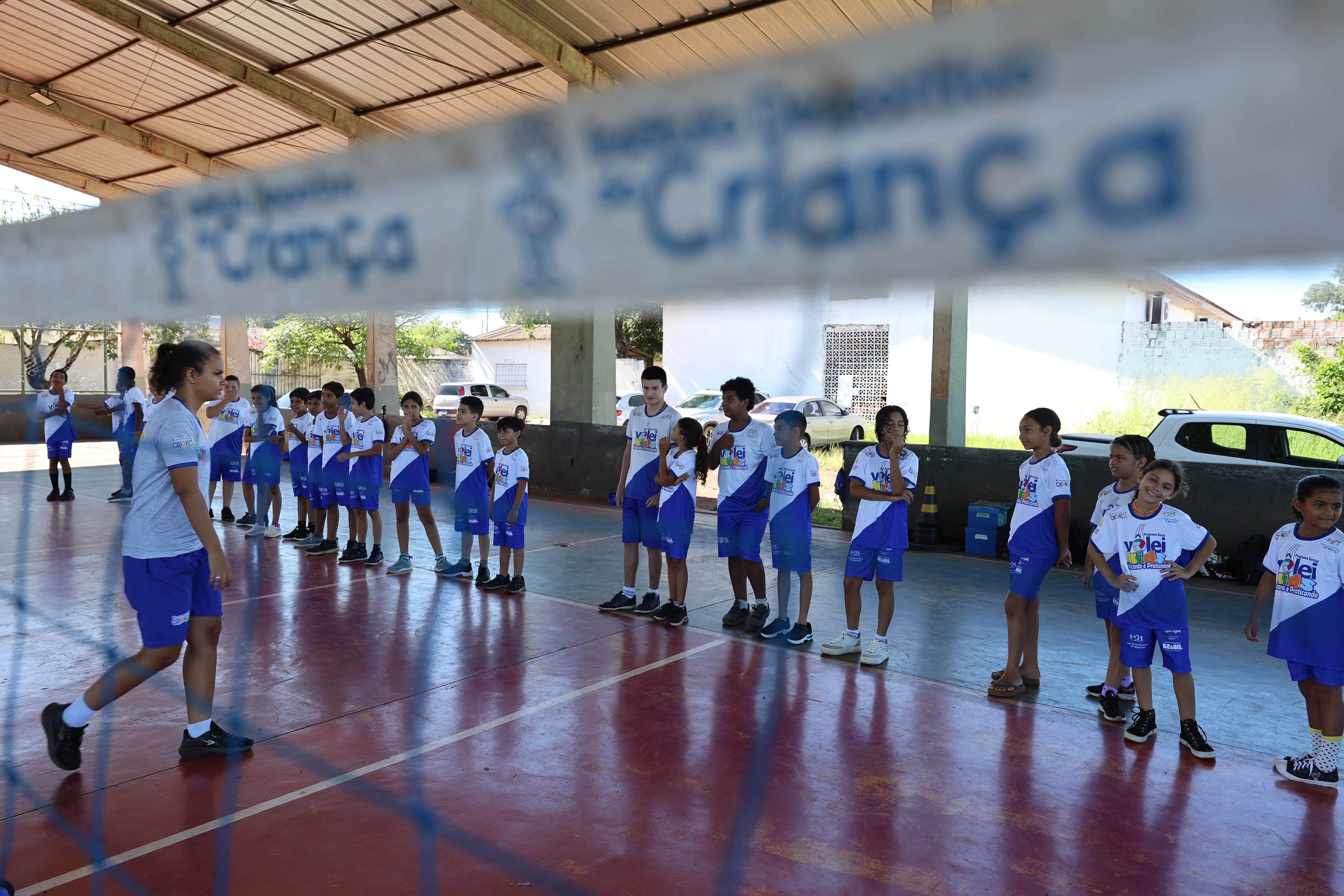 Grupo de crianças alinhadas em quadra, algumas com bola. Estão organizadas em fila, sob cobertura metálica.