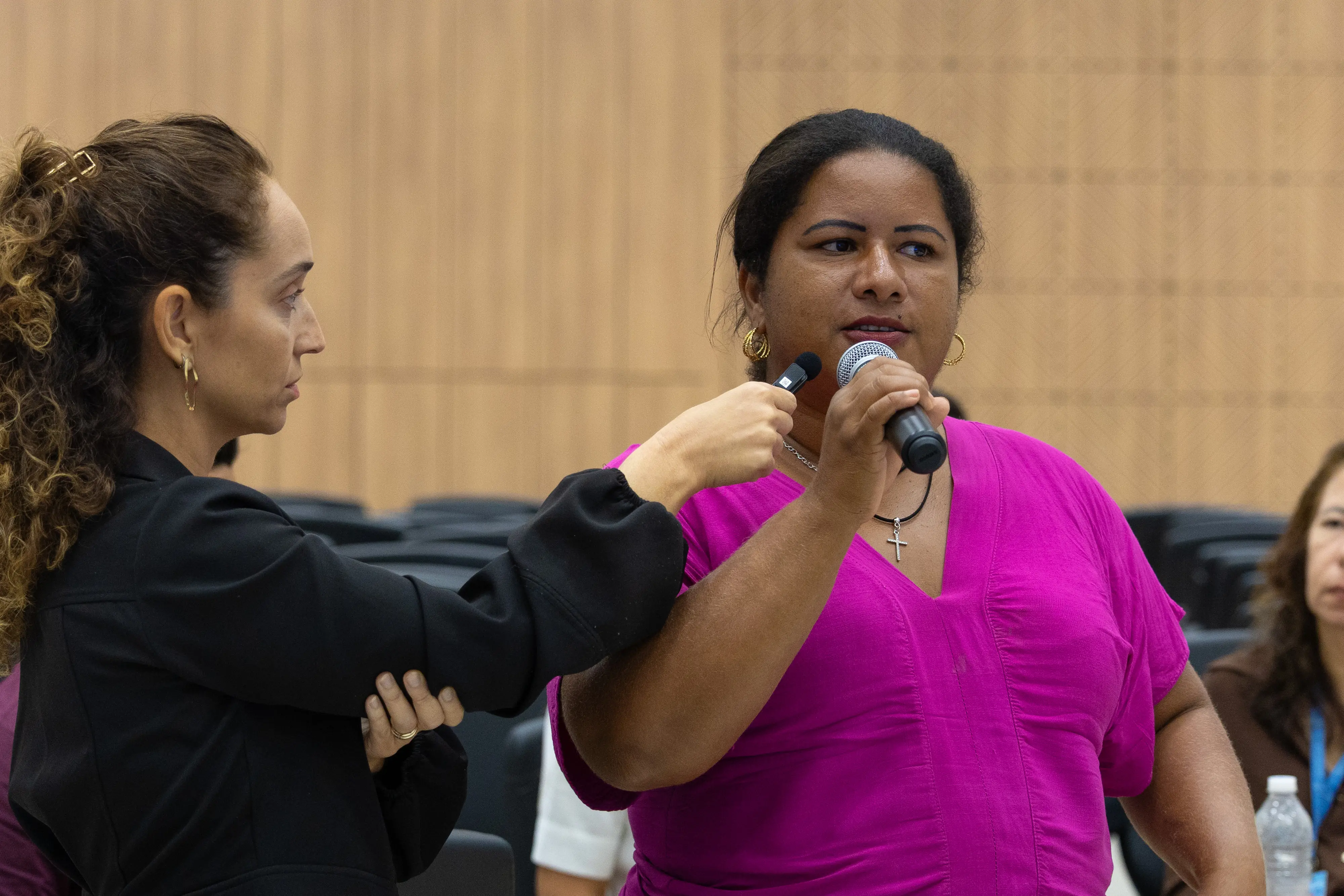 Mulher de blusa rosa fala ao microfone durante oficina. Participante ao lado segura gravador de áudio.