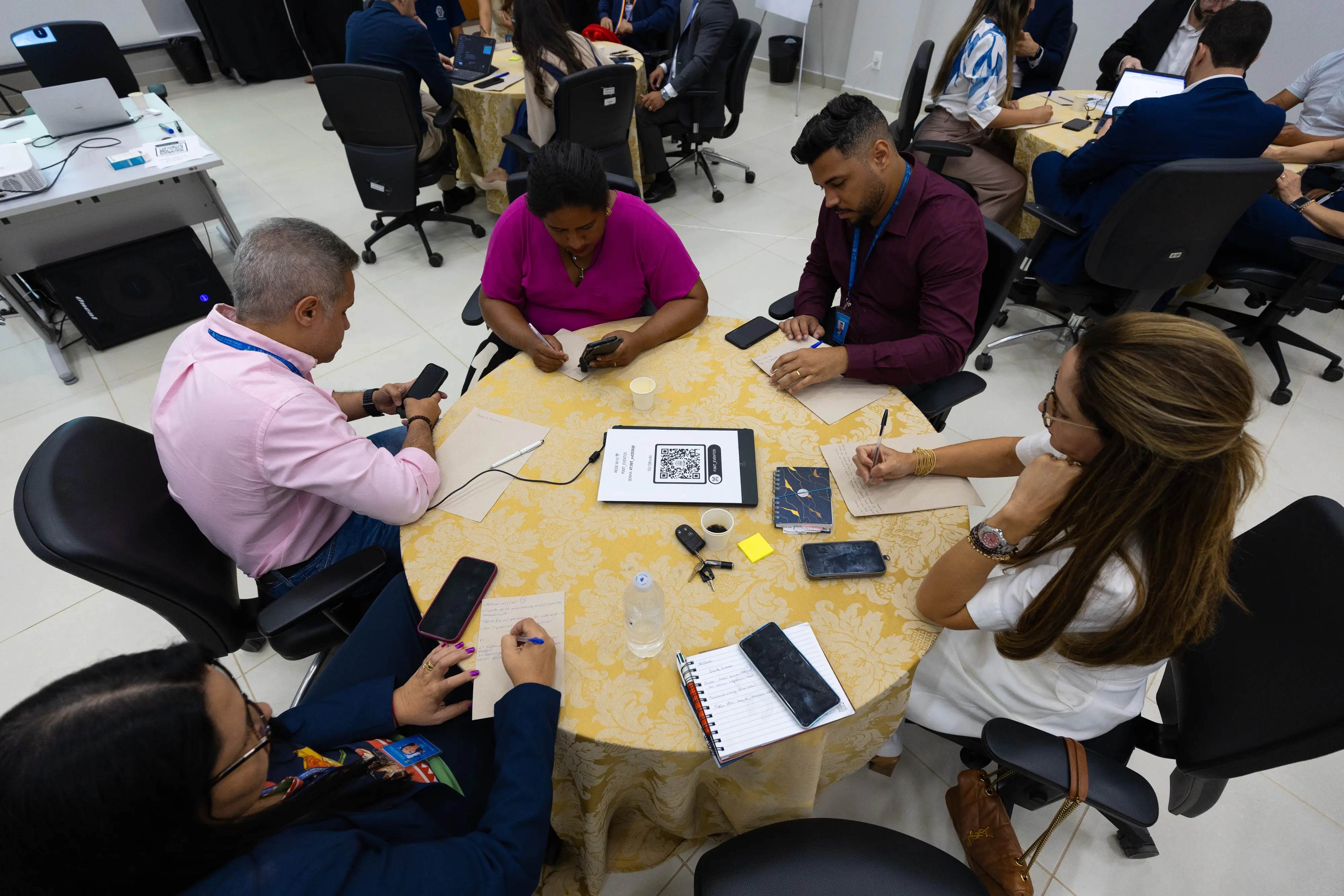 Participantes realizam atividade em grupo com cartões, celulares e anotações sobre a mesa durante oficina.