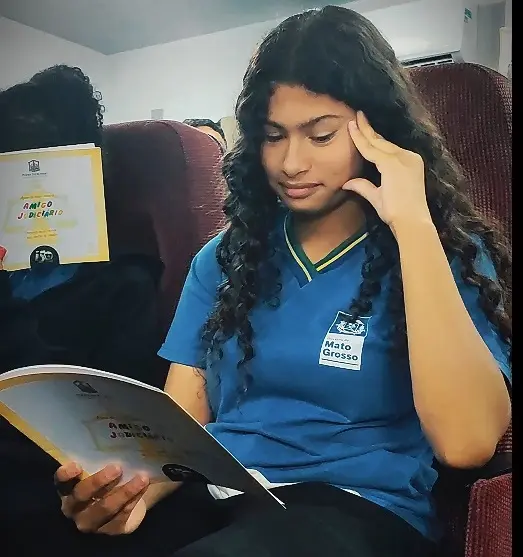 Uma menina está sentada lendo a cartilha do Nosso Judiciário. Ela usa uniforme azul, calça preta, tem cabelos longos, pretos, cacheados