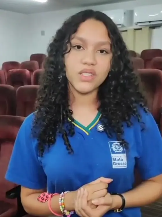 Estudantes com uniforme azul durante projeto Nosso Judiciário. Ela é uma adolescente branca, de cabelos com pridos cacheados, usa pulseiras e relógio