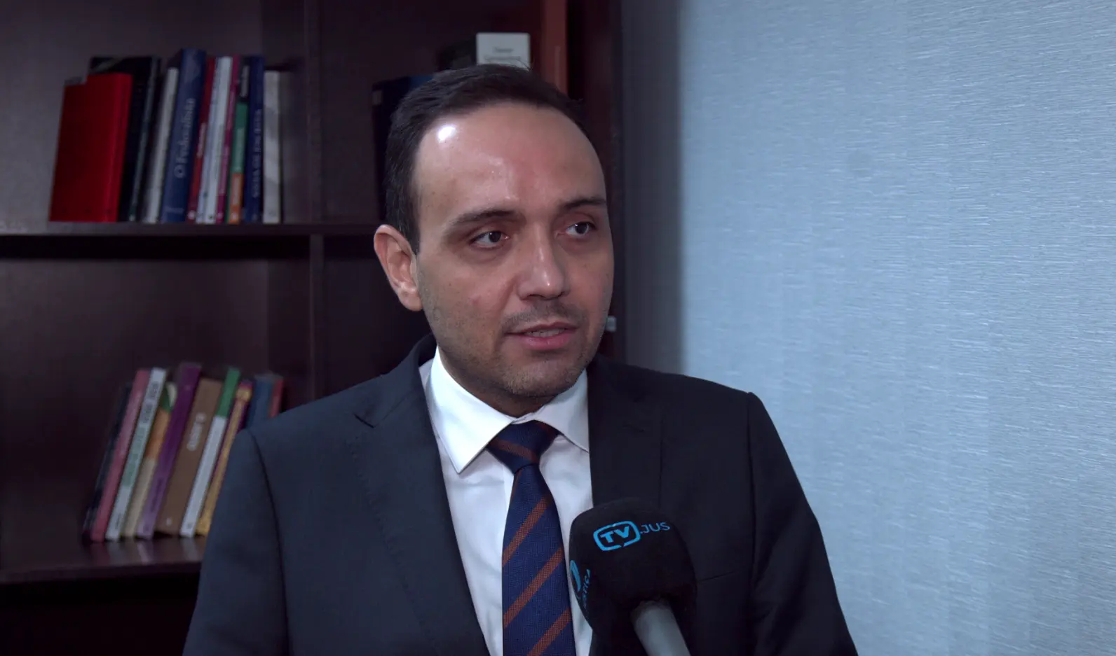 Juiz Gerardo Humberto concede entrevista à TV.Jus. ele é um homem de olhos e cabelos escuros, usando camisa branca, gravata listrada azul e marrom e terno cinza escuro. Atrás dele há uma estante com livros. 