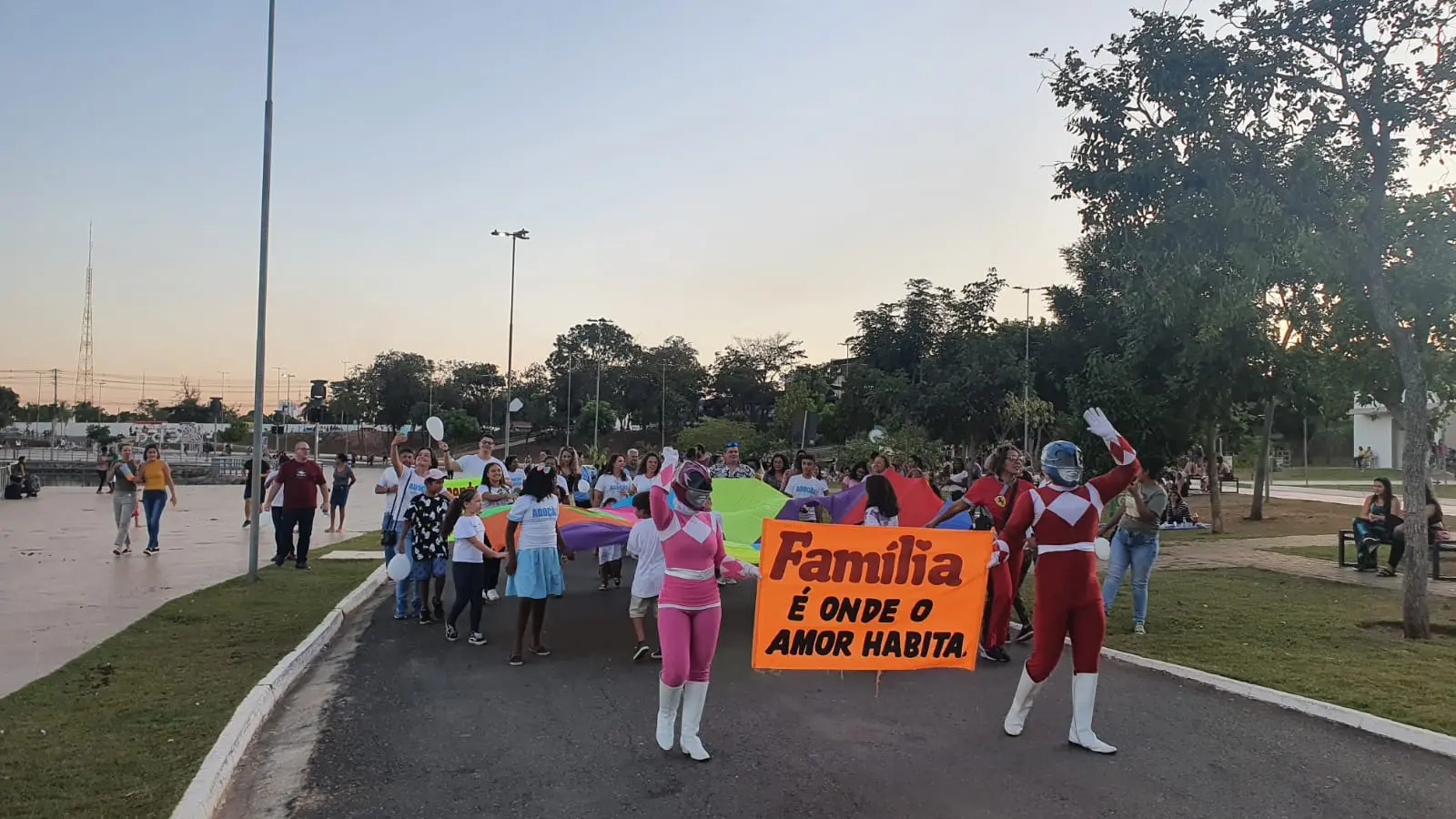 Imagem de edição anterior da Caminhada da Adoção. Adultos, crianças e duas pessoas fantasiadas de Power Rangers seguram uma grande bandeira colorida no Parque das Águas. As pessoas usam camisetas da Adoção e seguram balões.