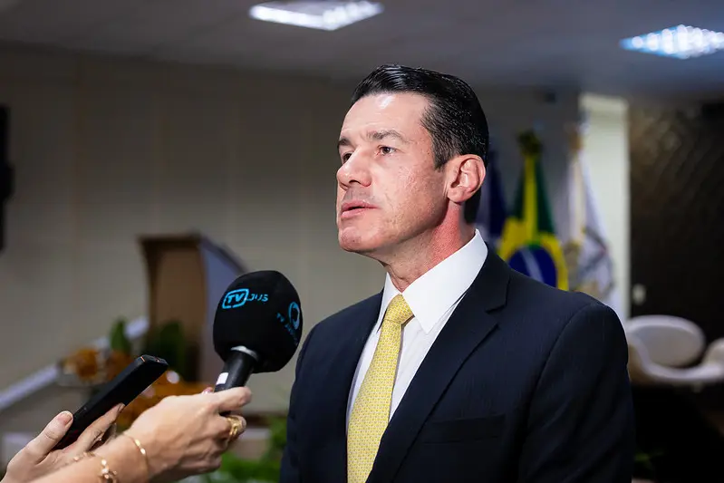 Foto horizontal que mostra o desembargador Rodrigo curvo, do busto pra cima, concedendo entrevista para a imprensa, em um auditório. Ele é um homem branco, alto, magro, de cabelos e olhos escuros, usando camisa branca, gravata amarelo e terno preto. 