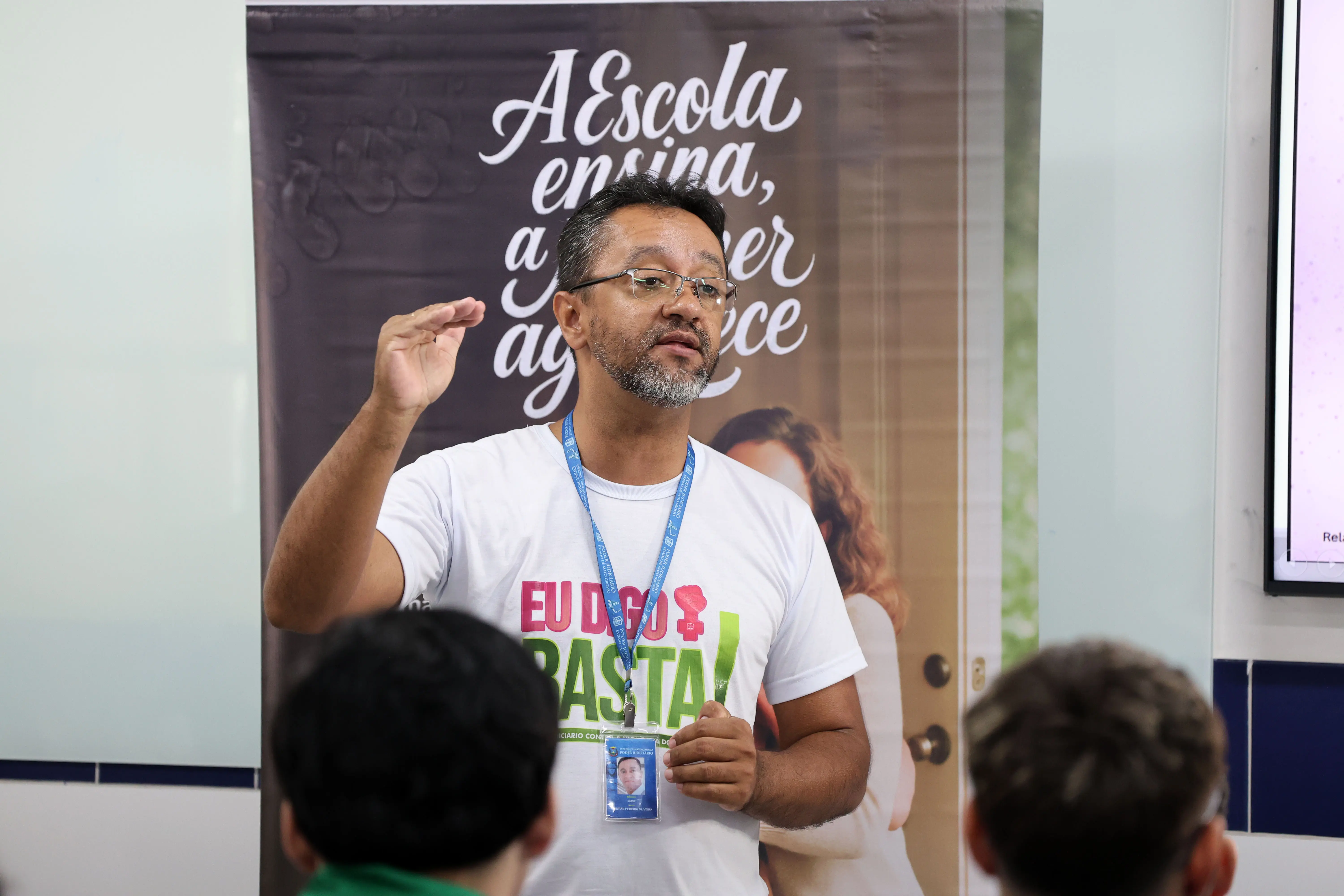 Plano médio de Cristian Pereira. Homem de óculos e barba curta, usando camiseta branca da campanha contra a violência. Ele está com a mão levantada, em meio a uma explicação, com um banner educativo ao fundo.