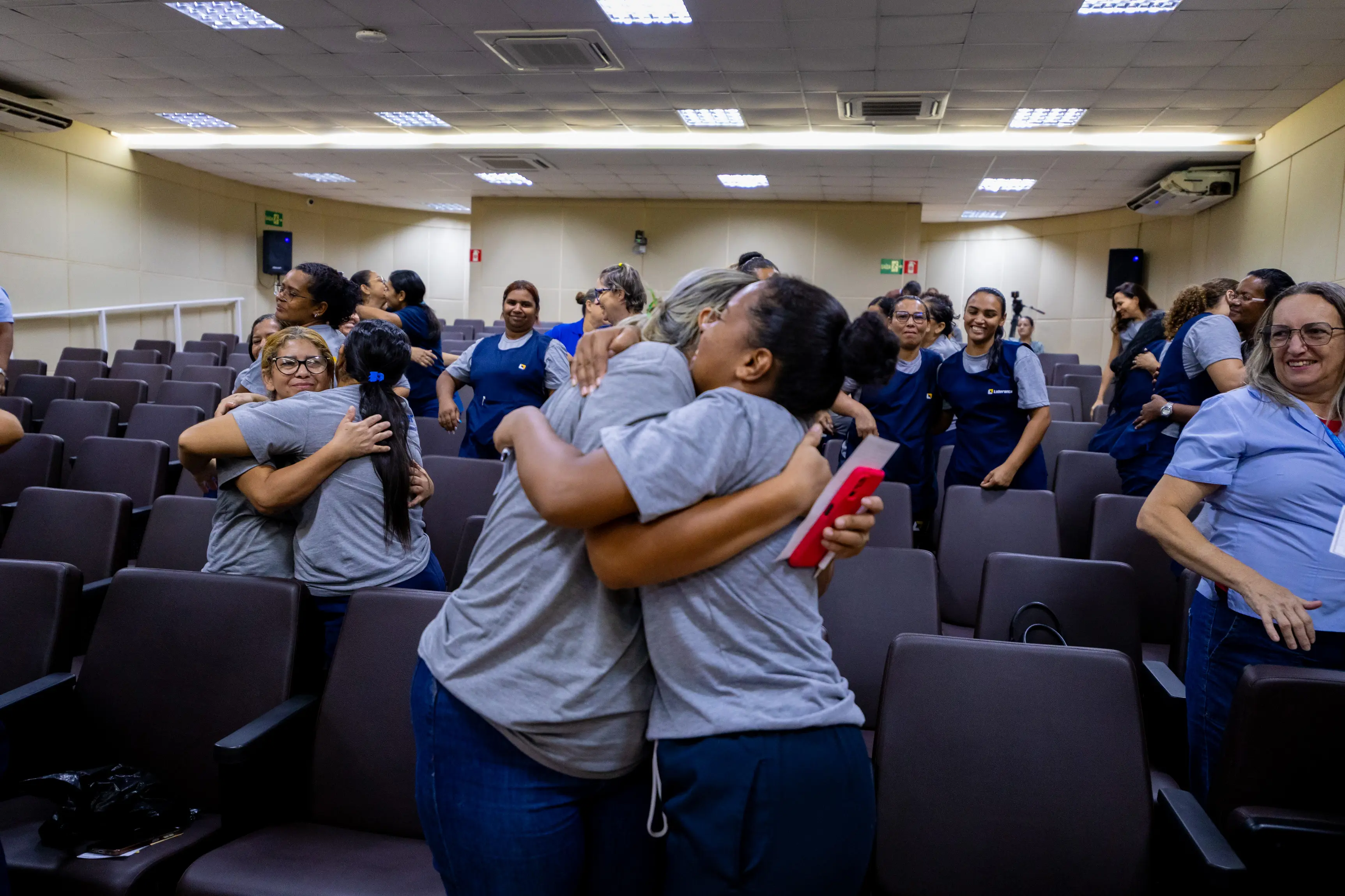 Participantes de um encontro institucional aparecem em pé entre as fileiras do auditório, formando duplas e trocando abraços durante dinâmica de integração. A maioria veste camisetas azuis, em clima de acolhimento.