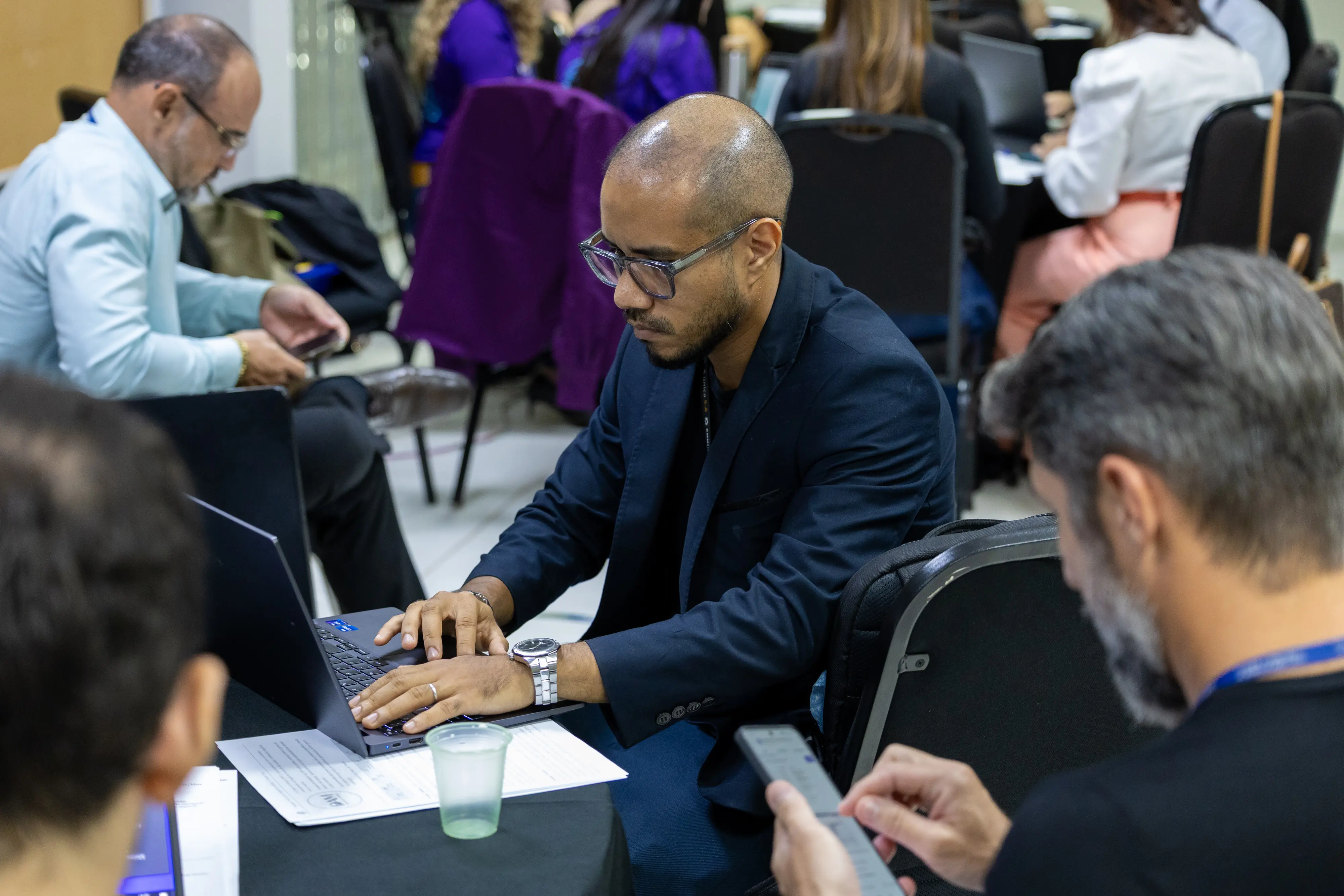Participante de blazer escuro digita no notebook com foco. Ao redor, colegas consultam celulares e papéis. O ambiente evidencia atividade simultânea entre dispositivos e discussão técnica.