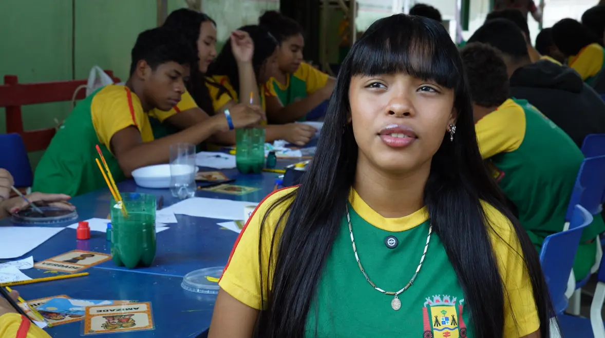 Jovem de franja e cabelos longos, veste camiseta verde e amarela com colares, está em pátio escolar cercada de colegas pintando à mesa.