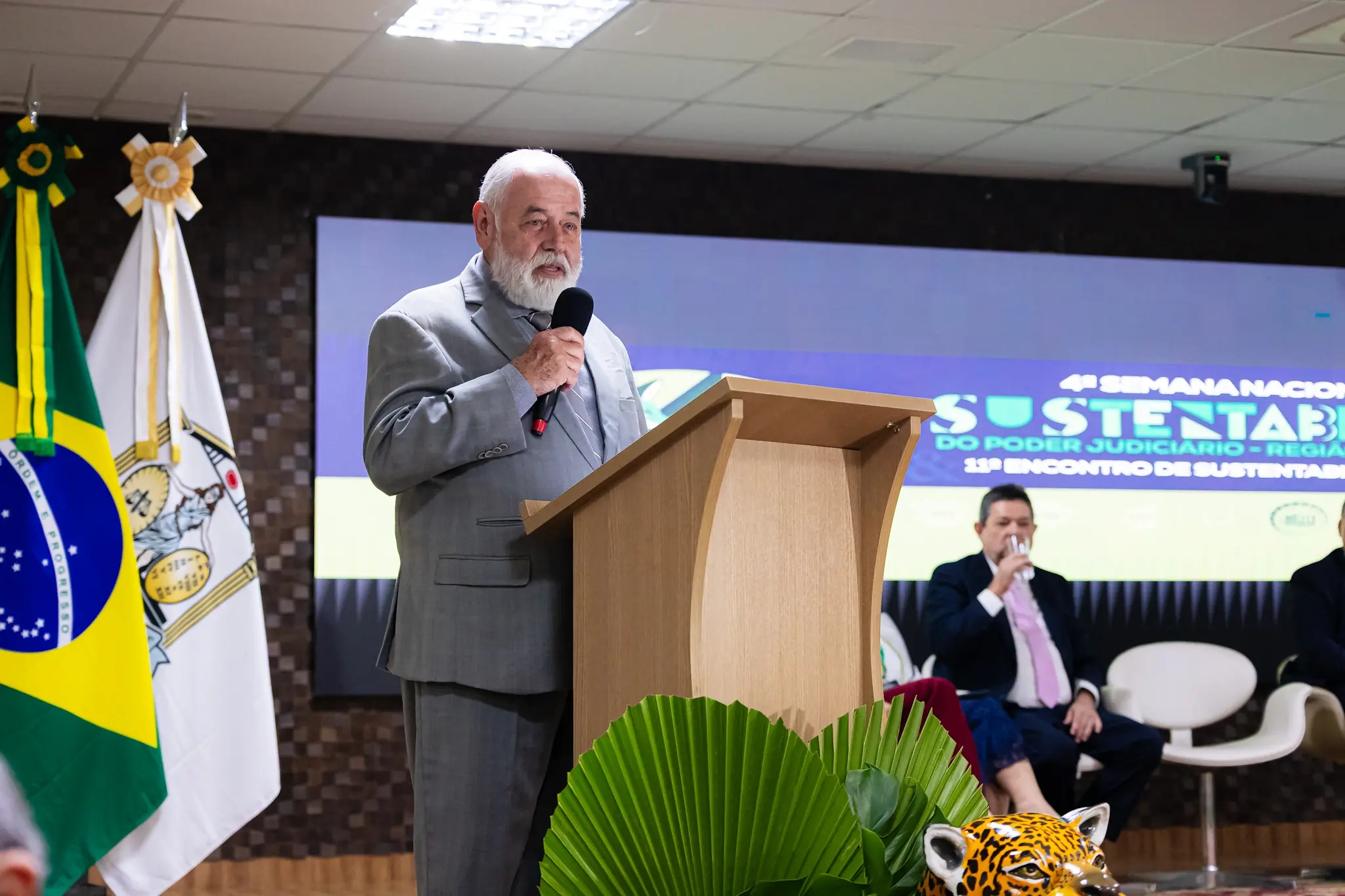 Homem idoso de barba e cabelos brancos, vestindo terno cinza, discursa em um púlpito de madeira. À esquerda, a bandeira do Brasil. Ao fundo, painel com o título do evento e pessoas dispostas no palco.