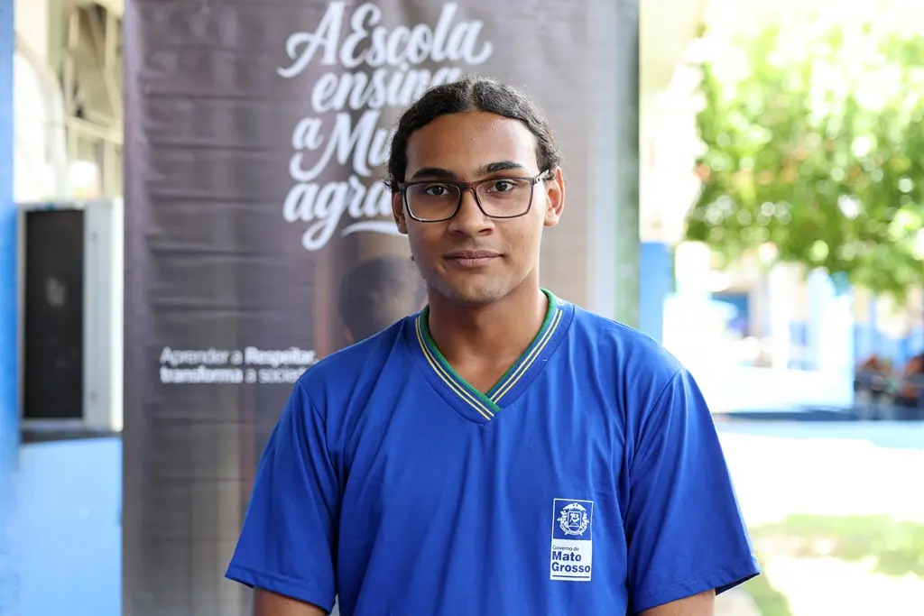 Foto horizontal em plano médio que mostra o estudante Lucas Daniel Queiroz posando para a foto, no pátio da escola. Ele é um adolescente negro, alto, de cabelo preso, olhos castanhos, usando óculos de grau e uniforme escolar azul marinho.