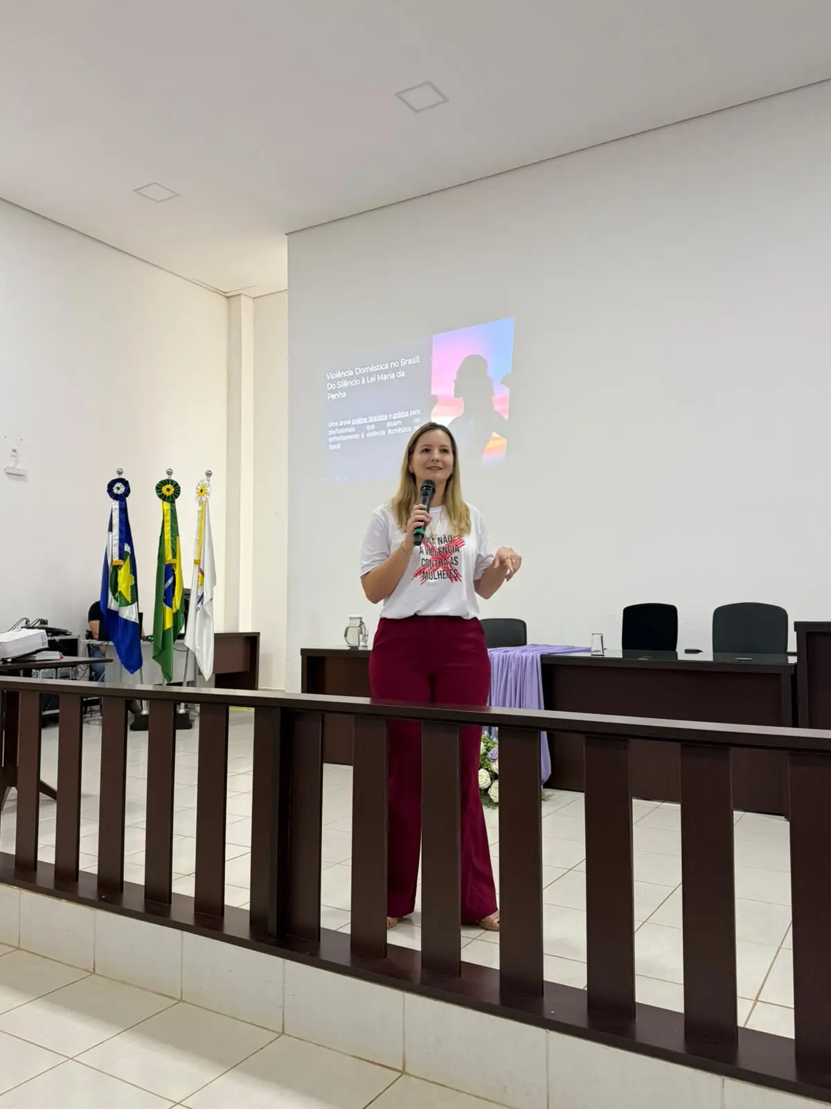 Foto vertical que mostra a juíza Djessica Küntzer em pé, falando ao microfone durante palestra no plenário do júri de Mirassol D'Oeste. Ela é uma mulher branca, loira, usando calça vermelha e camiseta branca com a estampa da frase "Diga não à violência contra a mulher" e um X vermelho em cima.