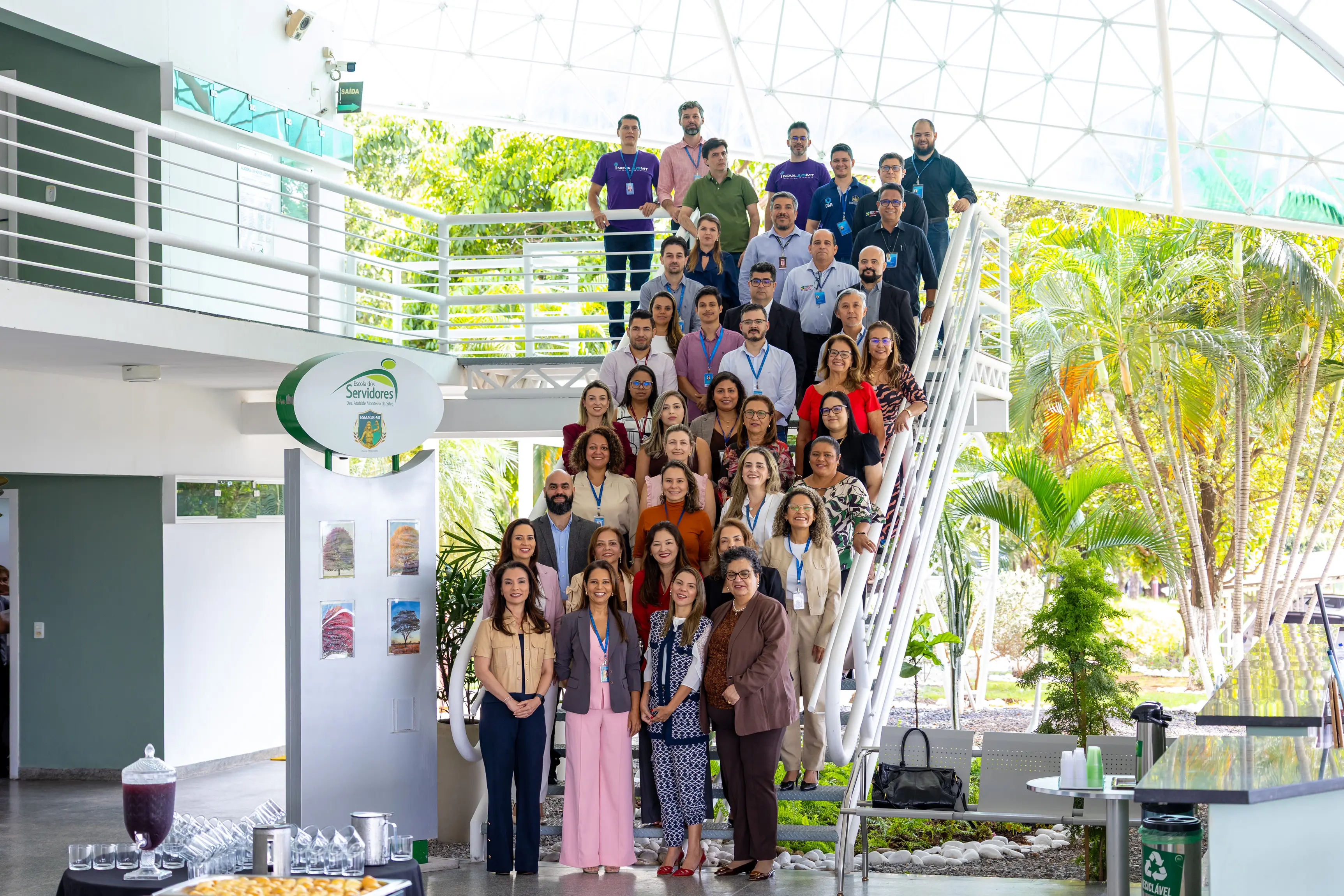 Foto em grupo com dezenas de pessoas posando em uma escadaria metálica branca em ambiente amplo e iluminado.