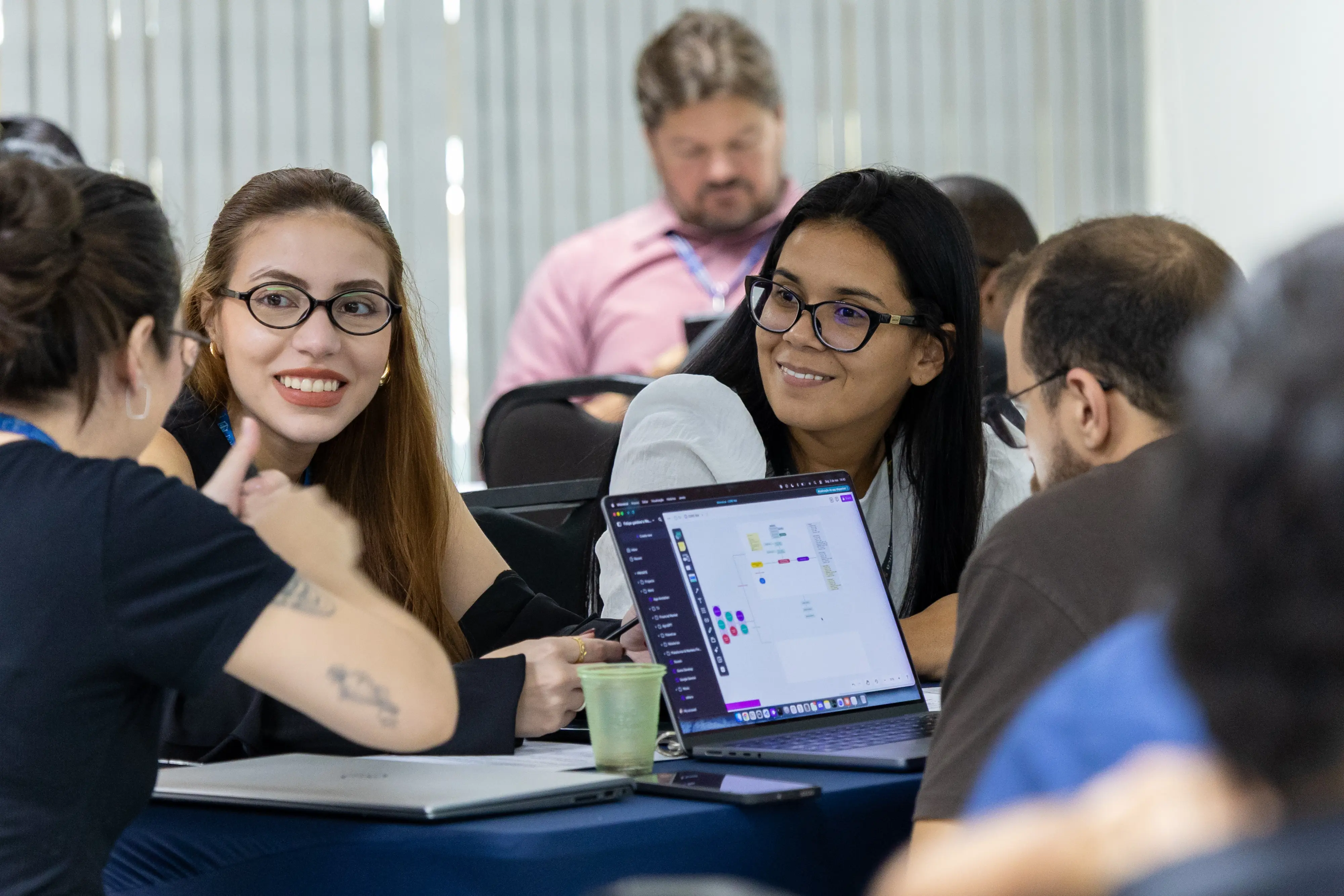 Duas mulheres trocam olhares e sorrisos durante conversa em grupo. Notebook aberto exibe painel digital, sugerindo análise de dados ou organização de ideias no exercício coletivo.
