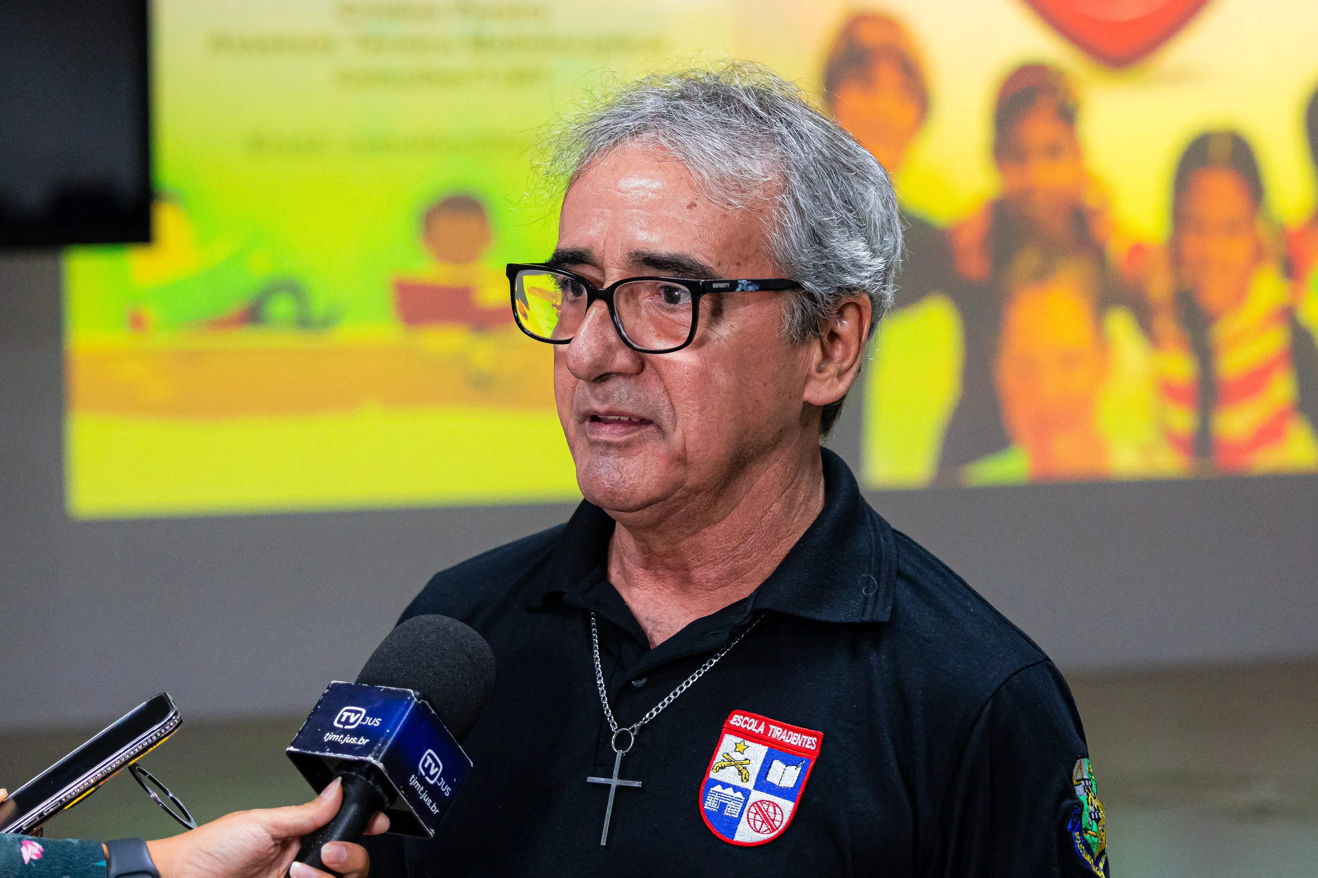 Homem de cabelos grisalhos, óculos e camisa preta com crucifixo, veste polo da Escola Tiradentes e é entrevistado por microfone da TV Jus.