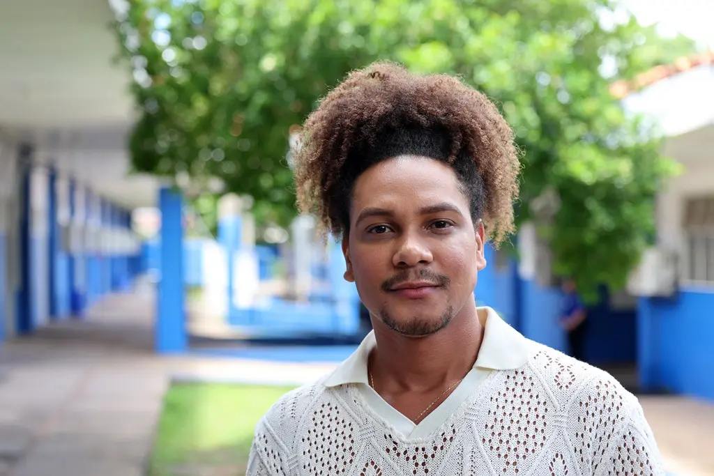 Foto horizontal em plano fechado que mostra o diretor da escola Cesário Neto, Wagner Mônantha, no pátio da unidade. Ele é um jovem negro, de olhos castanhos, cabelo crespo preso, barba, usando camiseta bege de renda.