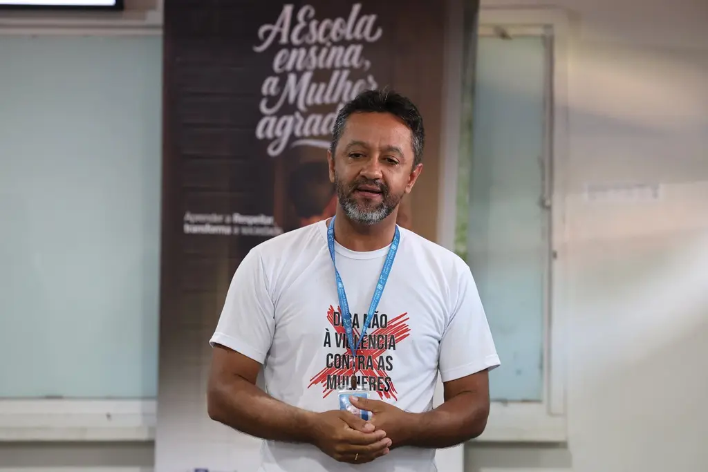 Foto horizontal em plano médio que mostra o assessor da Cemulher, Cristian Pereira, durante palestra em sala de aula. Ele é um homem negro, de cabelos castanhos, barba grisalha, usando camiseta branca com uma estampa onde se lê: Diga não à violência contra as mulheres.