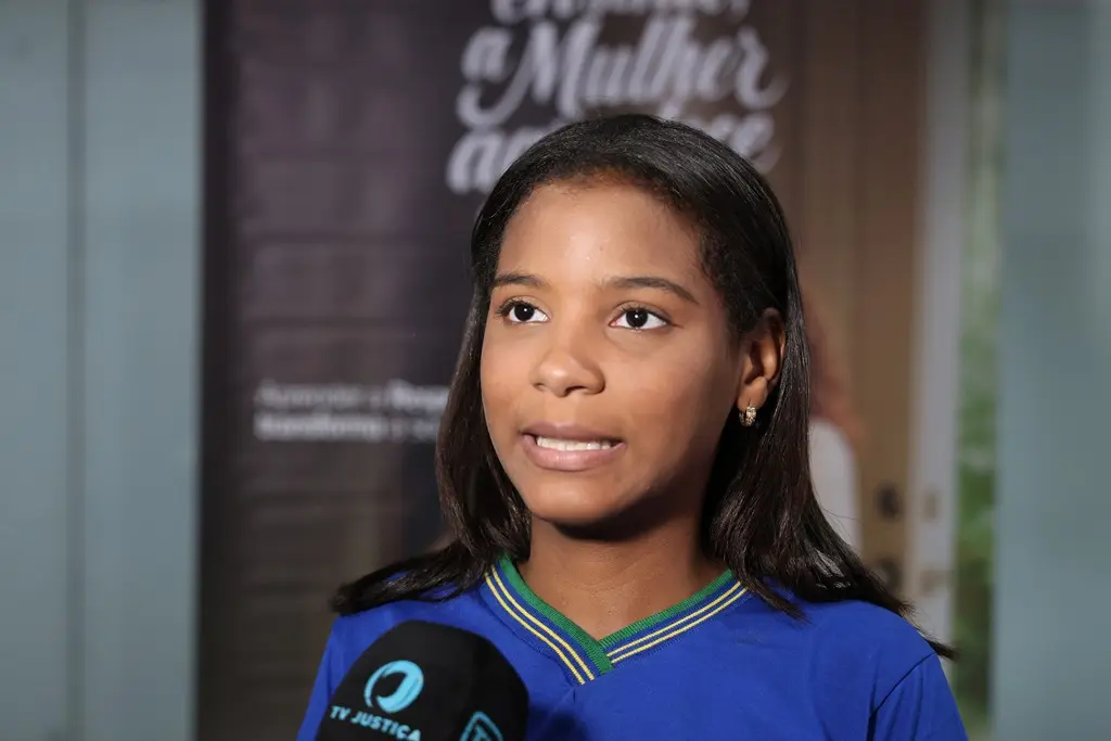 Foto horizontal em plano fechado que mostra a adolescente Ana Luiza Moraes durante entrevista à TV Justiça. Ela é negra, de cabelos pretos e alisados, olhos castanhos, usando uniforme escolar azul marinho. 