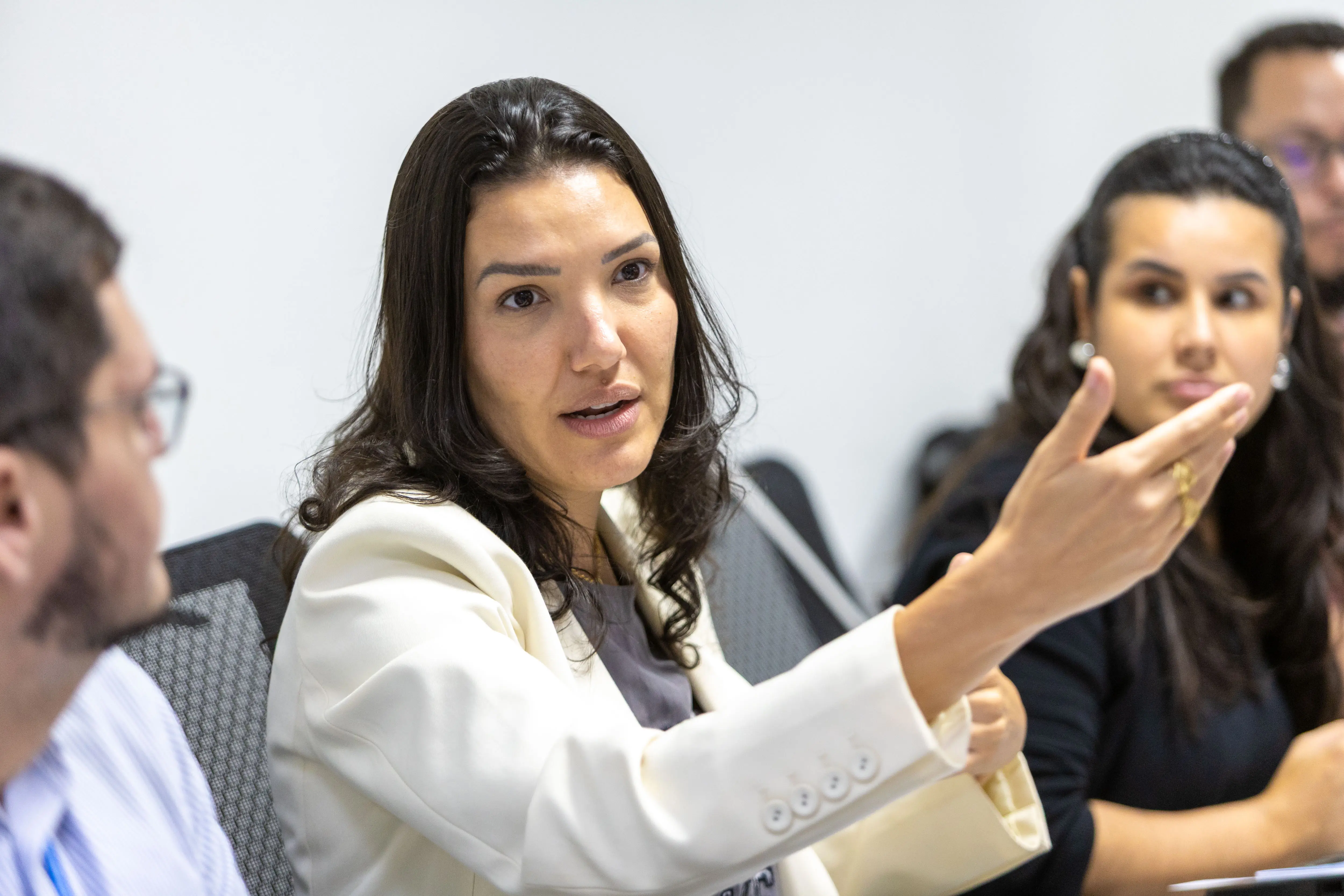 Mulher branca de cabelos escuros ondulados, usando blazer branco. Ela está sentada em uma mesa, gesticulando com a mão direita enquanto fala com alguém fora de foco. Ambiente de escritório moderno.