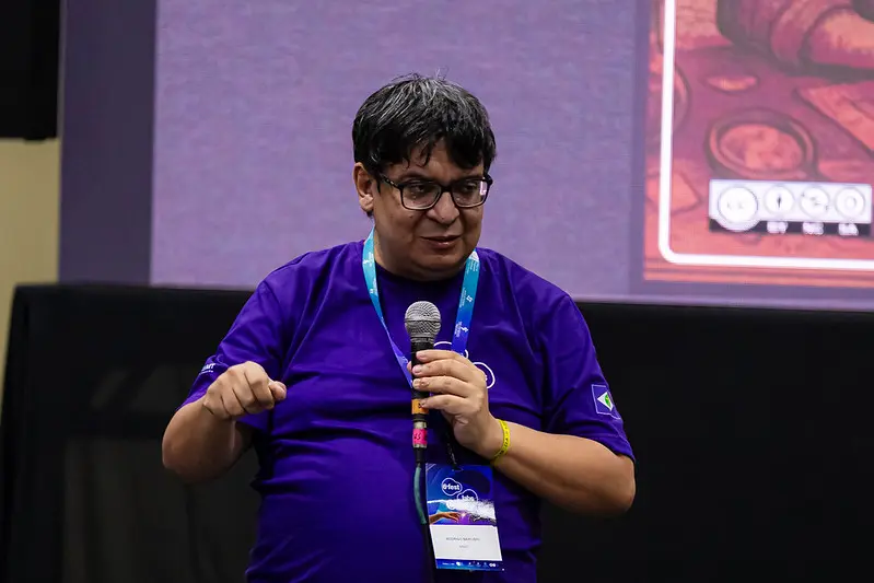 Foto horizontal que mostra Rodrigo Narciso conduzindo oficina no FestLabs. Ele aparece da cintura pra cima, gesticulando e falando ao microfone. Ele é um homem pardo, de cabelo preto, liso e preto, usando camiseta roxa do FestLabs, óculos de grau e crachá do evento. 