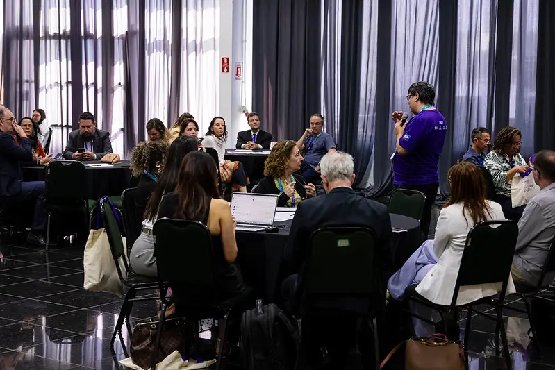 Foto horizontal em plano aberto que mostra uma sala com várias pessoas sentadas em volta de mesas redondas, participando de uma oficina do FestLabs Cuiabá. Em meio às mesas, aparece o instrutor, em pé, falando ao microfone, usando camiseta roxa.