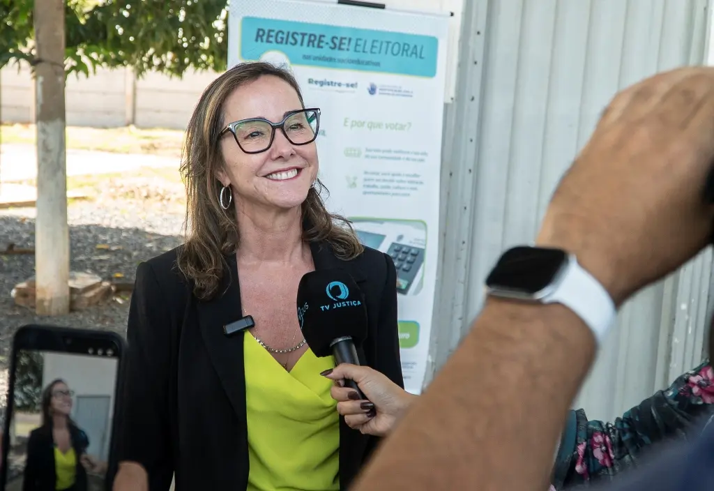 Foto horizontal que mostra a juíza Leilamar Rodrigues durante entrevista à TV Justiça. Ela é uma mulher branca, loira, usando blusa verde, blazer preto e óculos de grau.