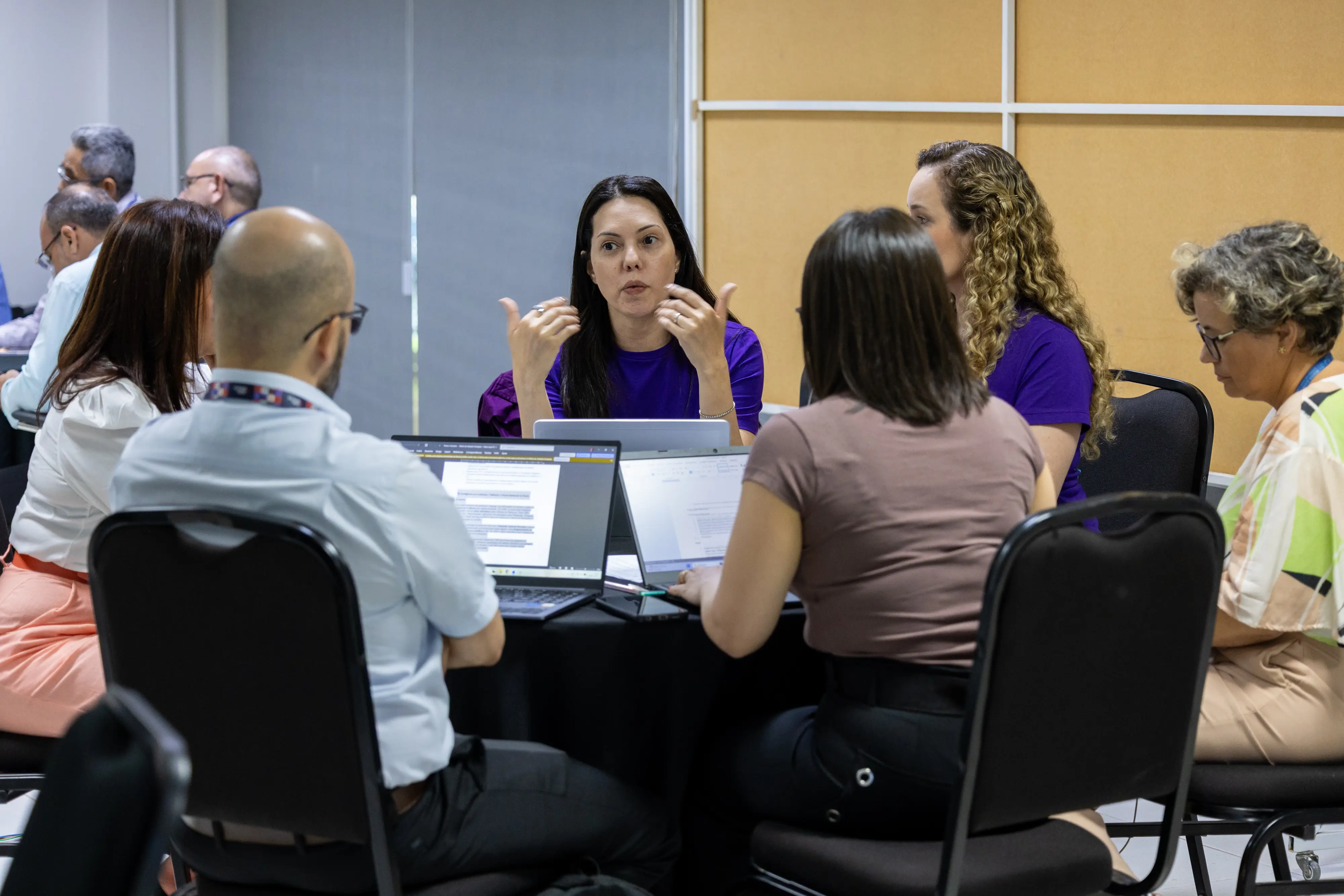 Mesa com equipe mista em debate. Mulher ao centro gesticula enquanto fala, liderando a discussão. Notebooks abertos e documentos impressos indicam fase de consolidação do trabalho.
