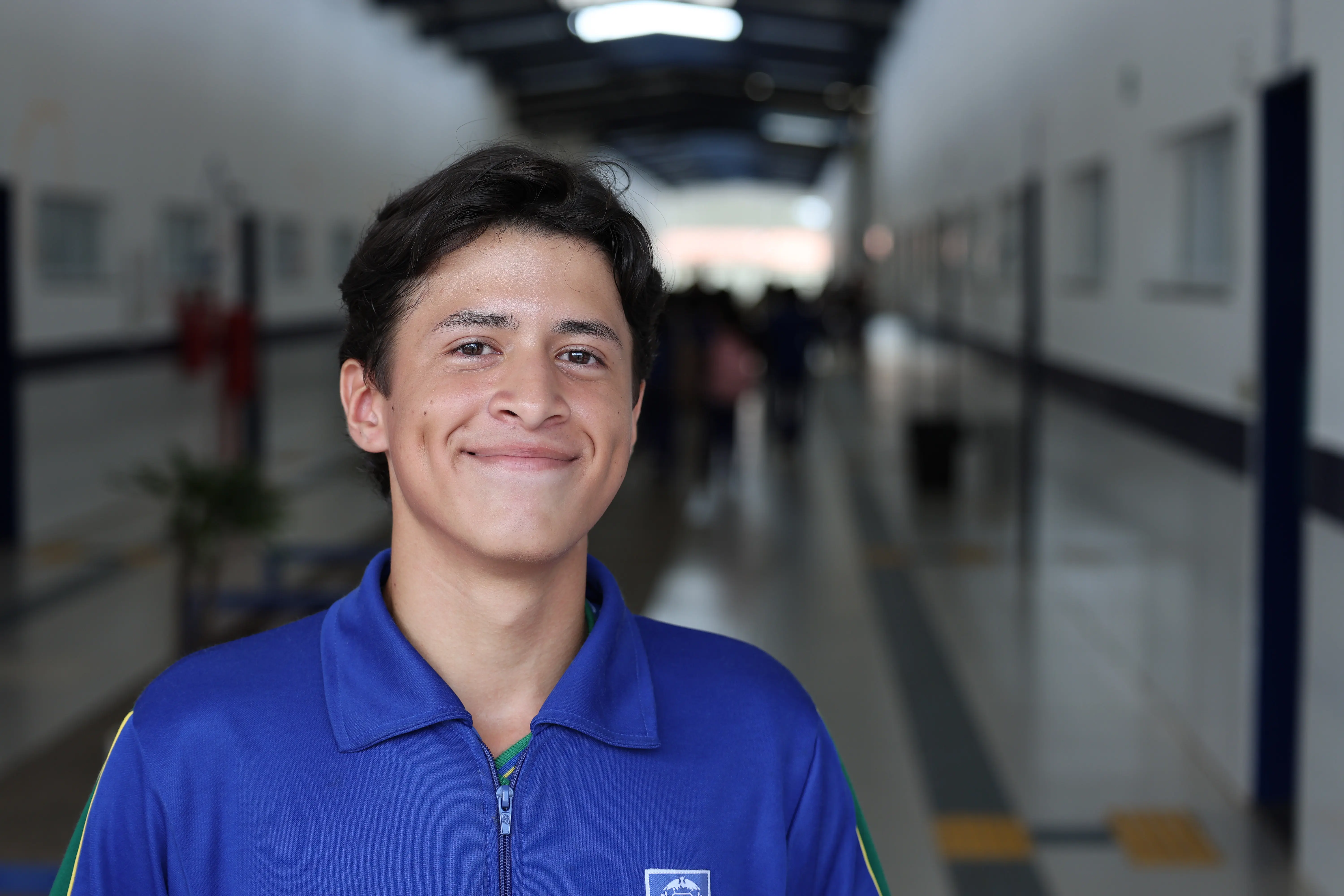 Retrato de Matheus Kaleby. Jovem sorridente de cabelos castanhos, vestindo o uniforme escolar azul com detalhes verdes. Ele está posicionado no centro de um corredor escolar que se estende ao fundo.