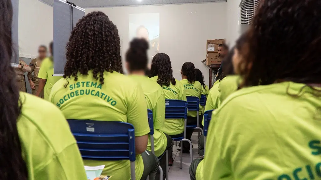 Foto horizontal que mostra várias meninas do sistema socioeducativo sentadas, assistindo a uma videoaula sobre as eleições. Elas estão de costas para a foto, todas usam camiseta verde neon do sistema socioeducativo.