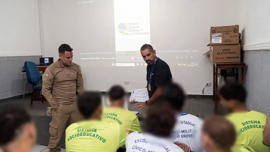 Foto horizontal que mostra uma sala cheia de adolescentes do sistema socioeducativo sentados, participando de uma atividade da Justiça eleitoral. Na frente deles, há um agente socioeducativo e um servidor da Justiça Eleitoral, que fala para eles. 