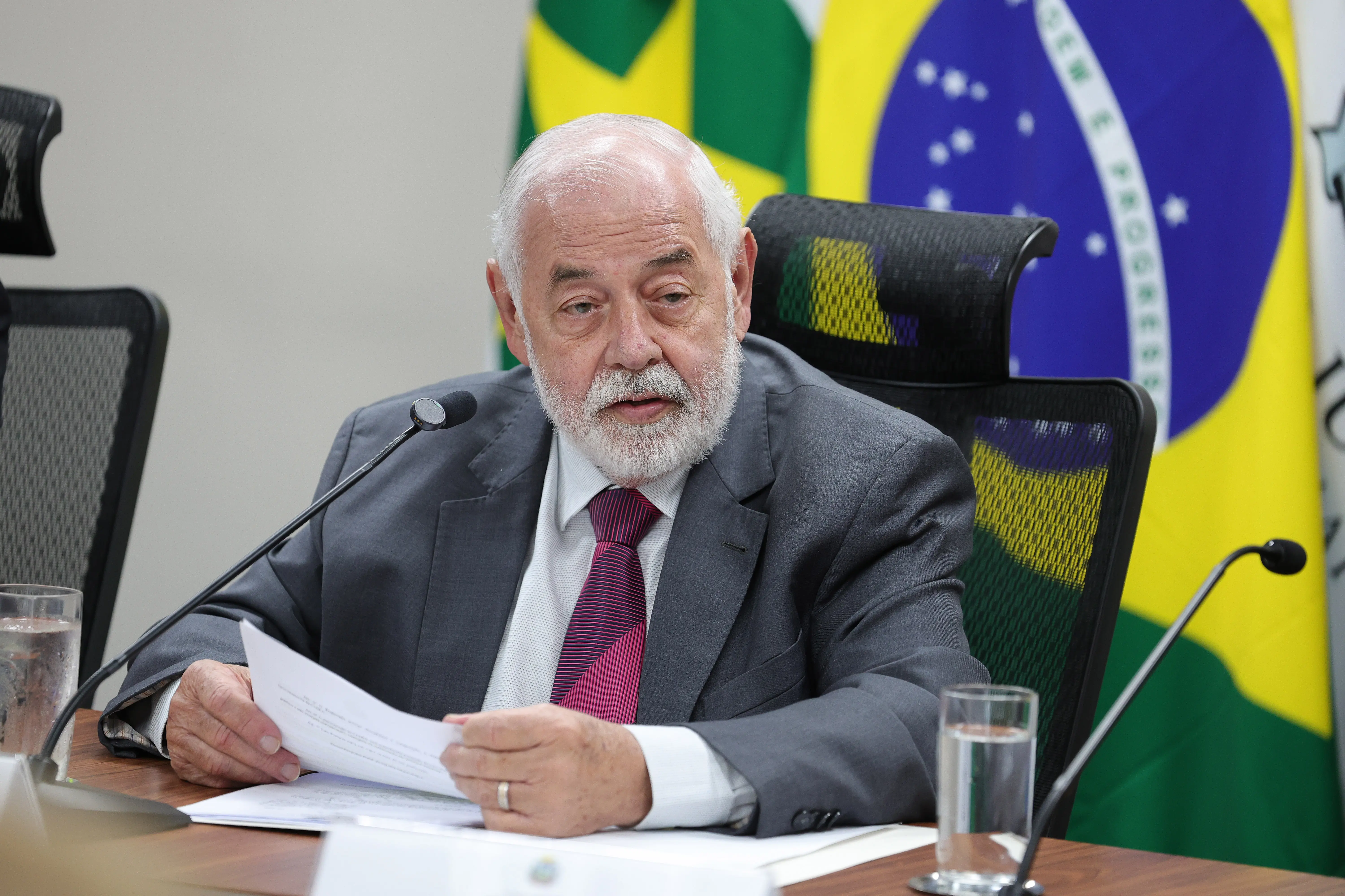 Plano médio de um homem idoso de cabelo e barba brancos, vestindo terno cinza, camisa branca e gravata vinho. Ele segura papéis na mesa e está diante de um microfone. Ao fundo, vê-se a bandeira do Brasil.
