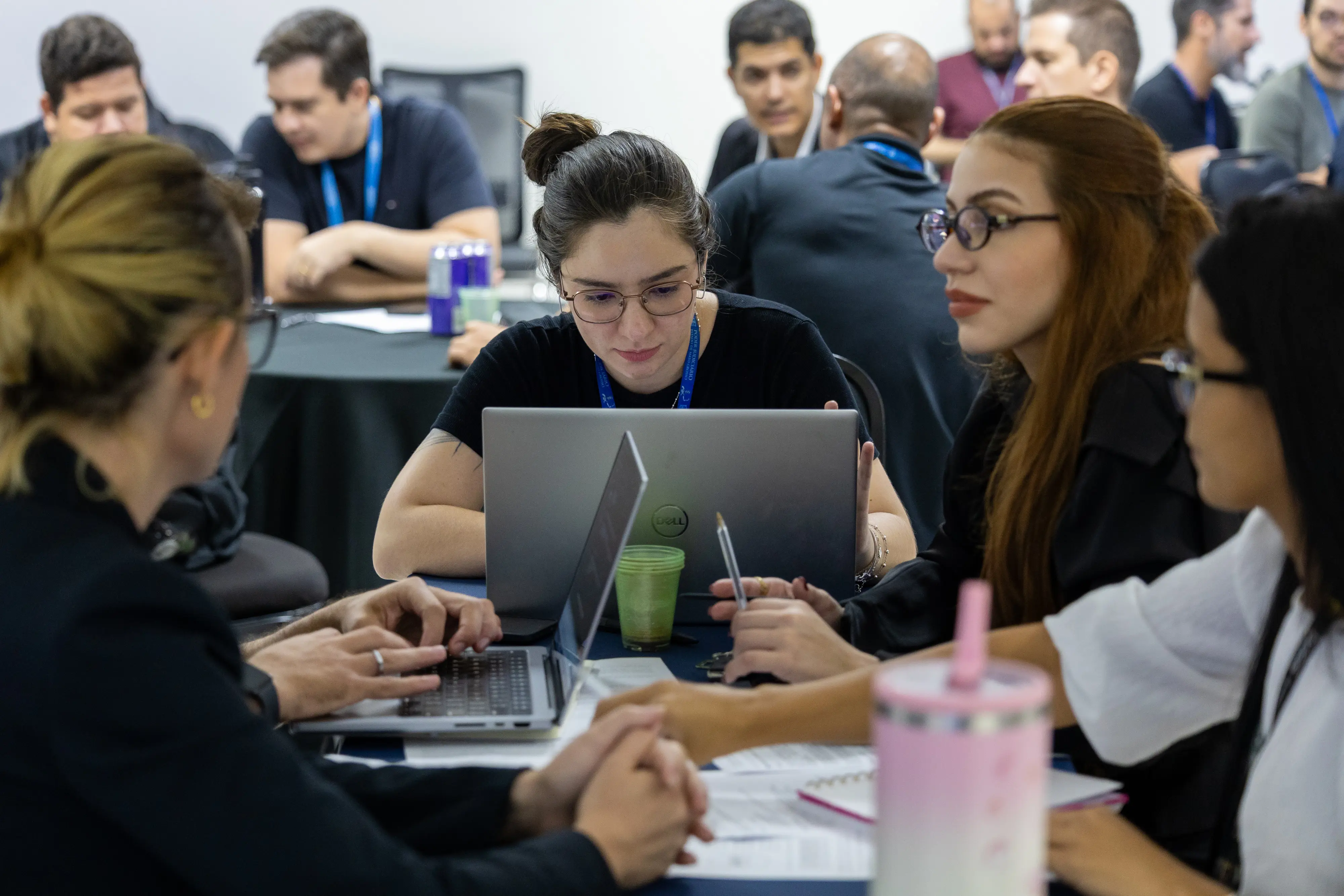 Grupo feminino reunido em mesa redonda trabalha em notebooks. Uma participante central observa a tela com atenção enquanto colegas digitam e anotam. Copos plásticos e materiais apoiam a atividade colaborativa.