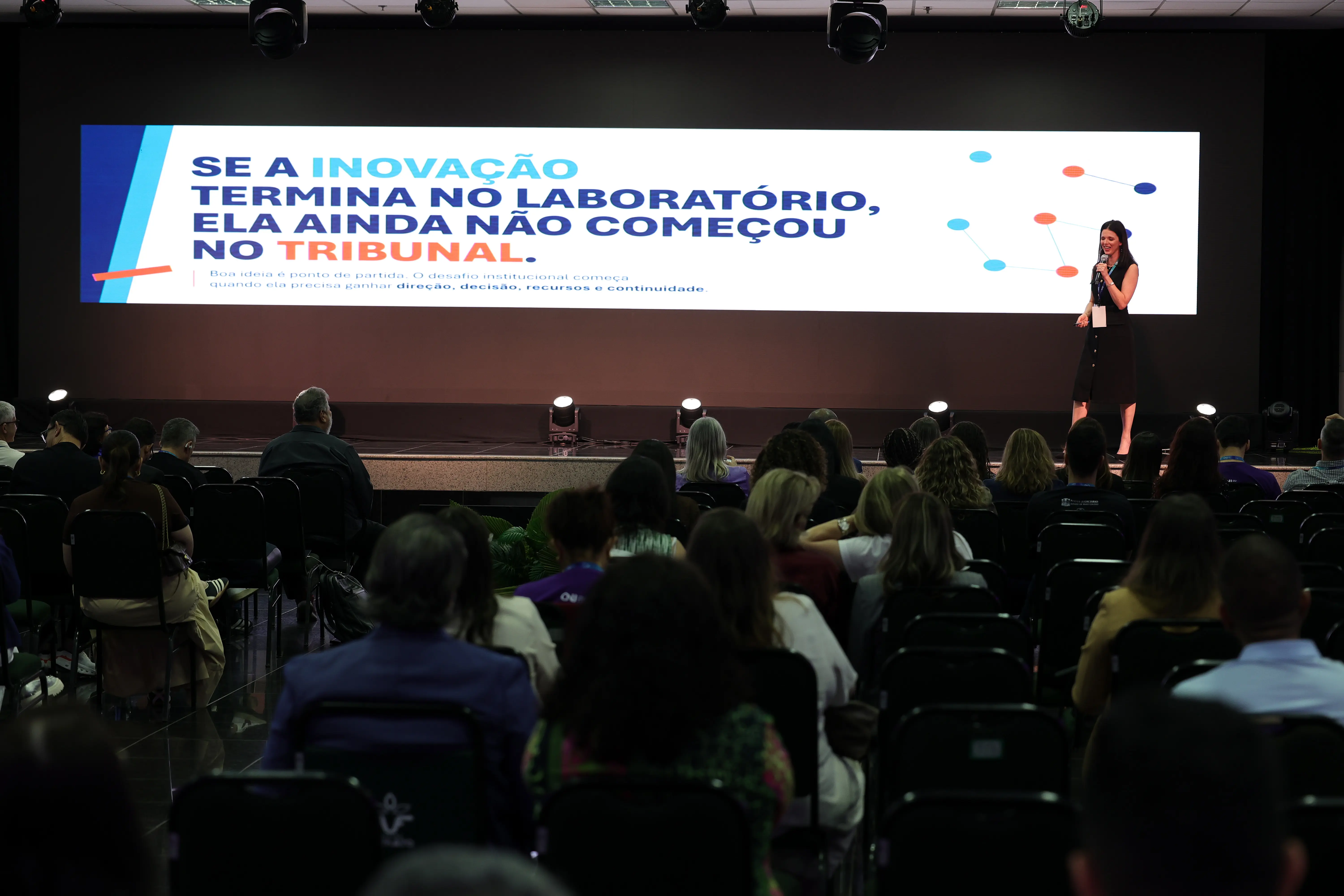 Vista do fundo do auditório lotado, mostrando a plateia sentada em cadeiras pretas. No palco, uma palestrante de vestido preto segura um microfone ao lado de um telão panorâmico que exibe a frase "SE A INOVAÇÃO TERMINA NO LABORATÓRIO, ELA AINDA NÃO COMEÇOU NO TRIBUNAL."