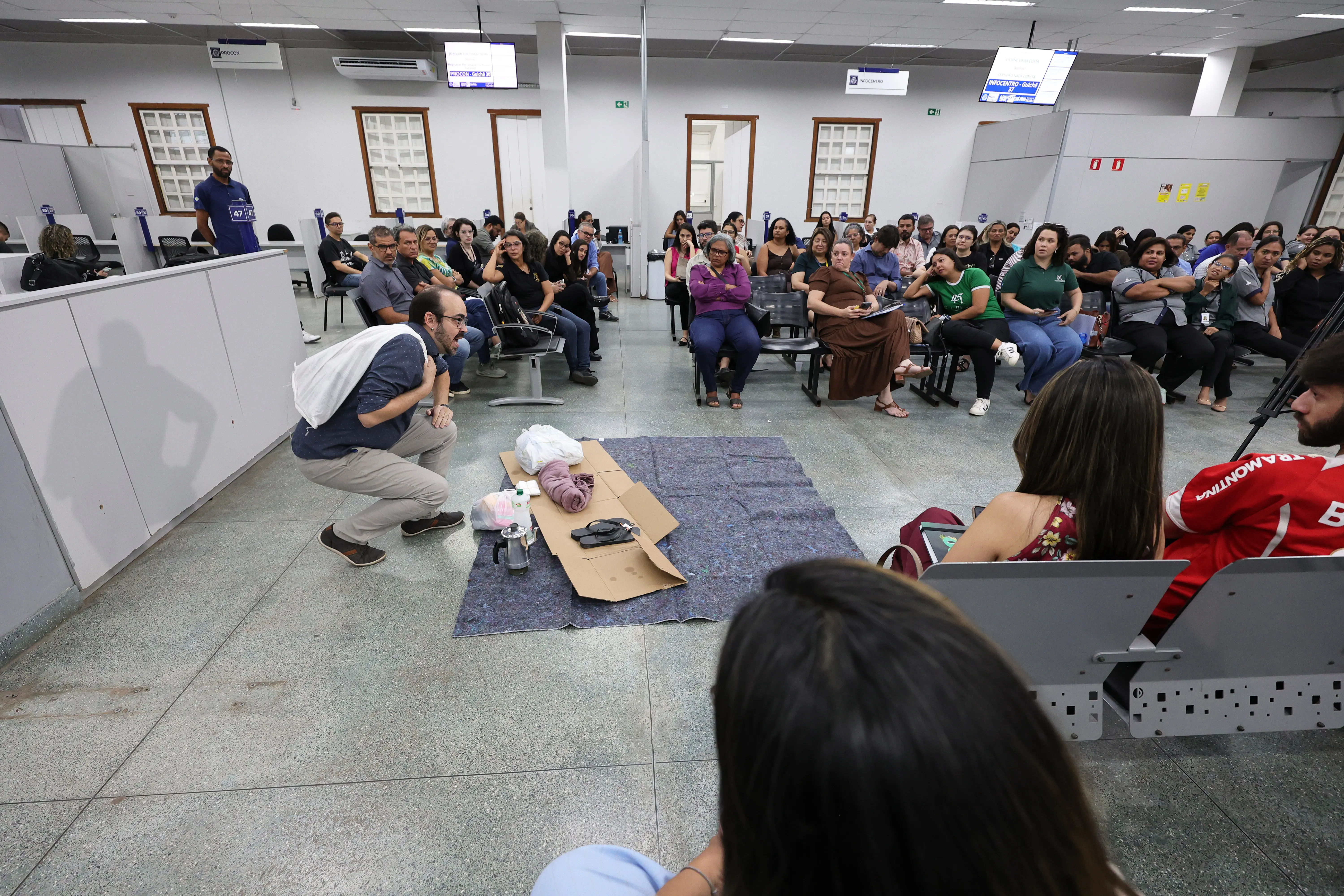 Homem de camisa azul e calça bege encena situação de rua, agachado ao lado de colchonete, cobertor, sacolas e pertences, diante de plateia atenta sentada em cadeiras.