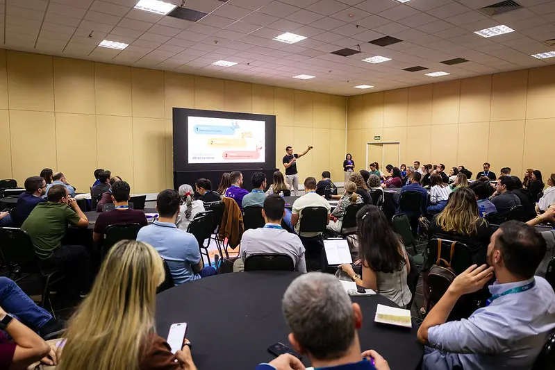 Foto horizontal em plano aberto que mostra uma grande sala com dezenas de pessoas sentadas em volta de mesas redondas, participando de uma oficina do FestLabs. Ao fundo, aparece um telão e o instrutor, em pé, falando ao microfone.
