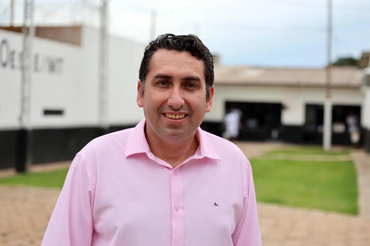 Retrato em primeiro plano de um homem sorridente de pele clara e cabelos escuros, vestindo uma camisa social rosa claro. O fundo mostra o pátio externo de uma unidade prisional de Mato Grosso.