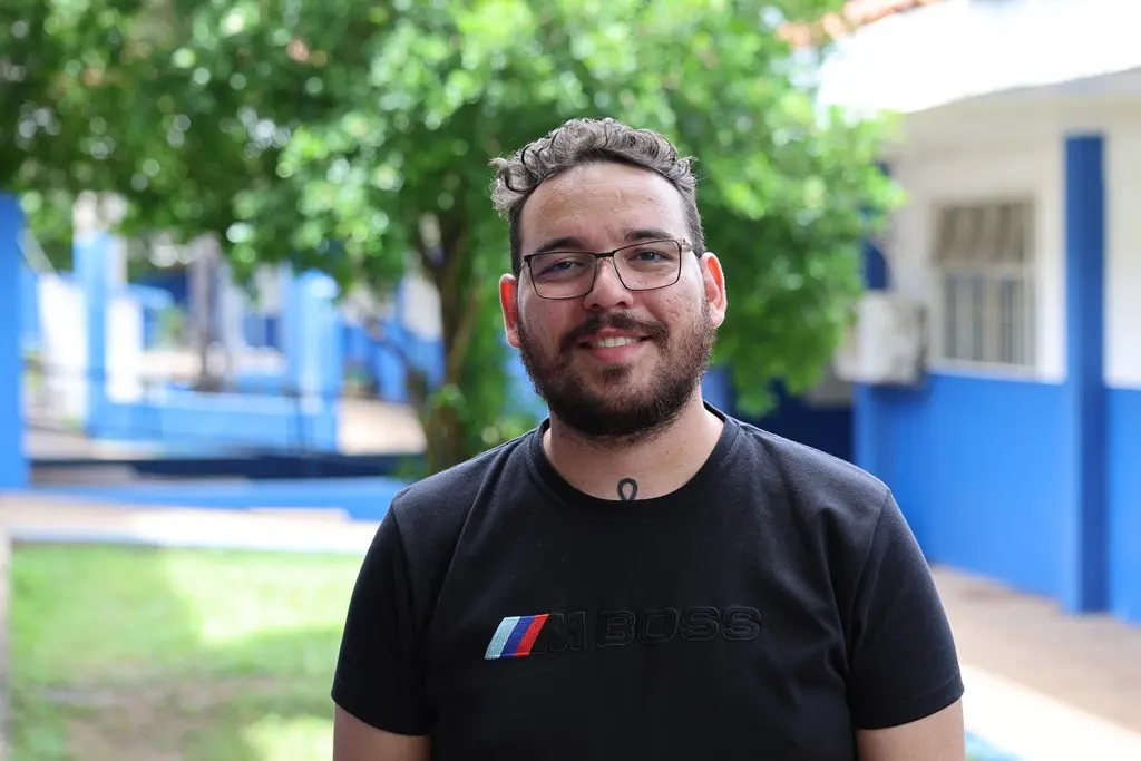 Foto horizontal em plano médio que mostra o psicólogo da Escola Cesário Neto, Lucas Patrick, sorrindo para a foto, no pátio da unidade. Ele é um jovem branco, de cabelo e barba castanhos claros, usando camiseta preta e óculos de grau. 