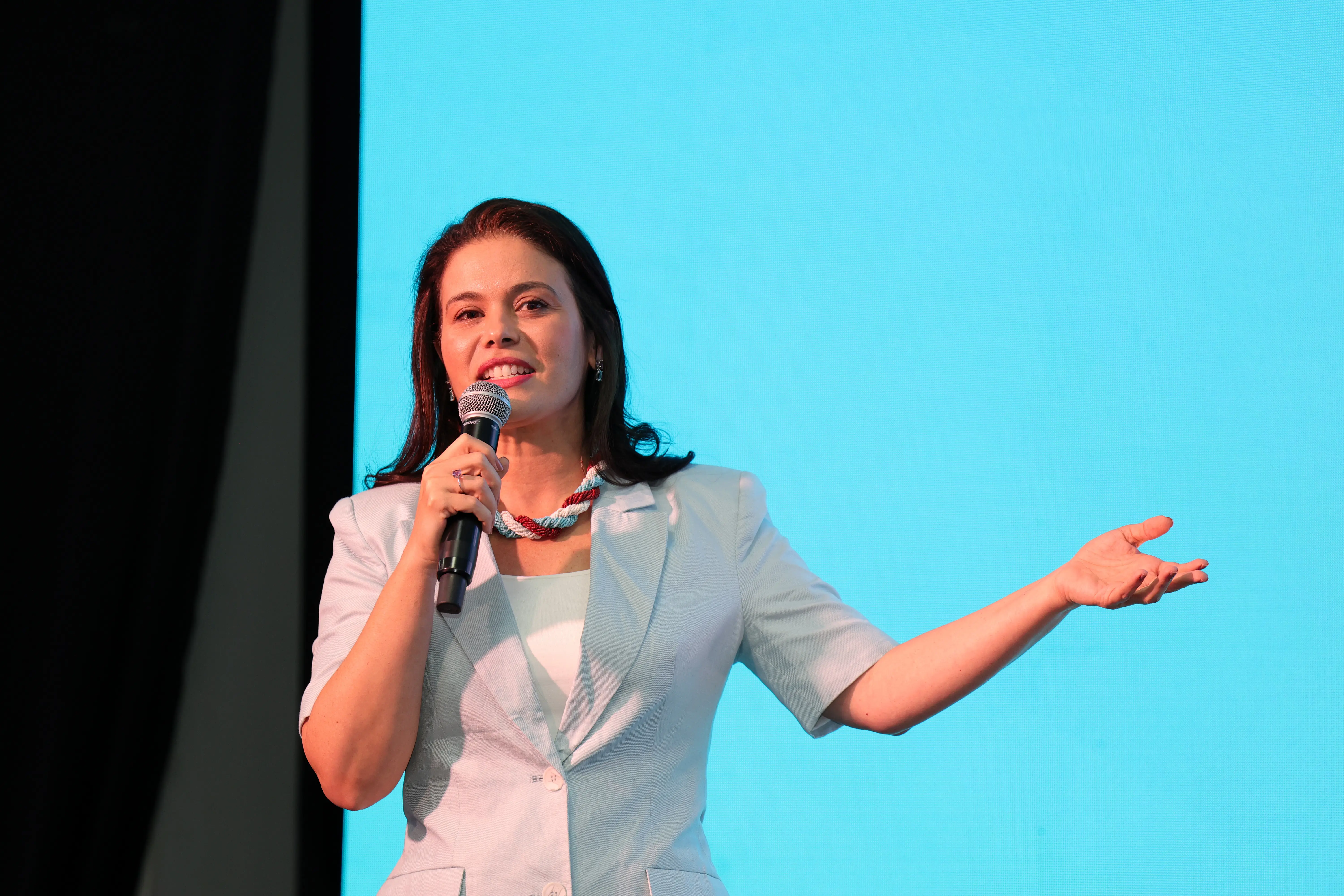 Juíza Luciana discursa ao microfone, de blazer azul-claro e colar étnico, gesticula com a mão direita diante de telão azul.