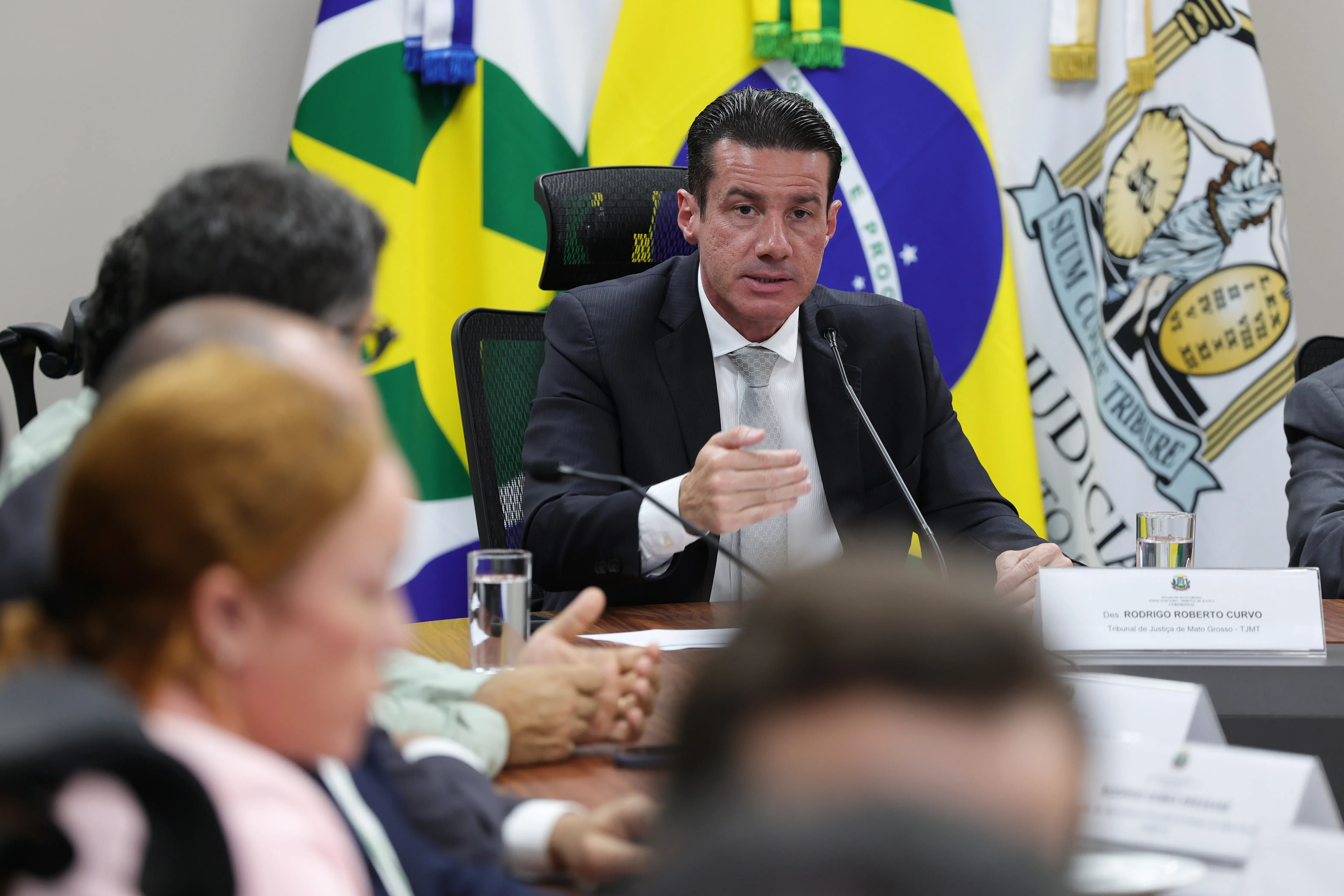 Plano médio de um homem de terno escuro, camisa branca e gravata cinza, falando com a mão direita estendida. Ele está sentado à mesa de reunião, com microfones e a bandeira do Brasil ao fundo.