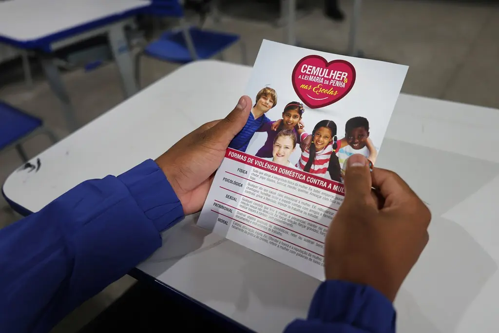 Foto horizontal em plano fechado que mostra as mãos de um estudante segurando um panfleto da Cemulher sobre os tipos de violência contra a mulher. 