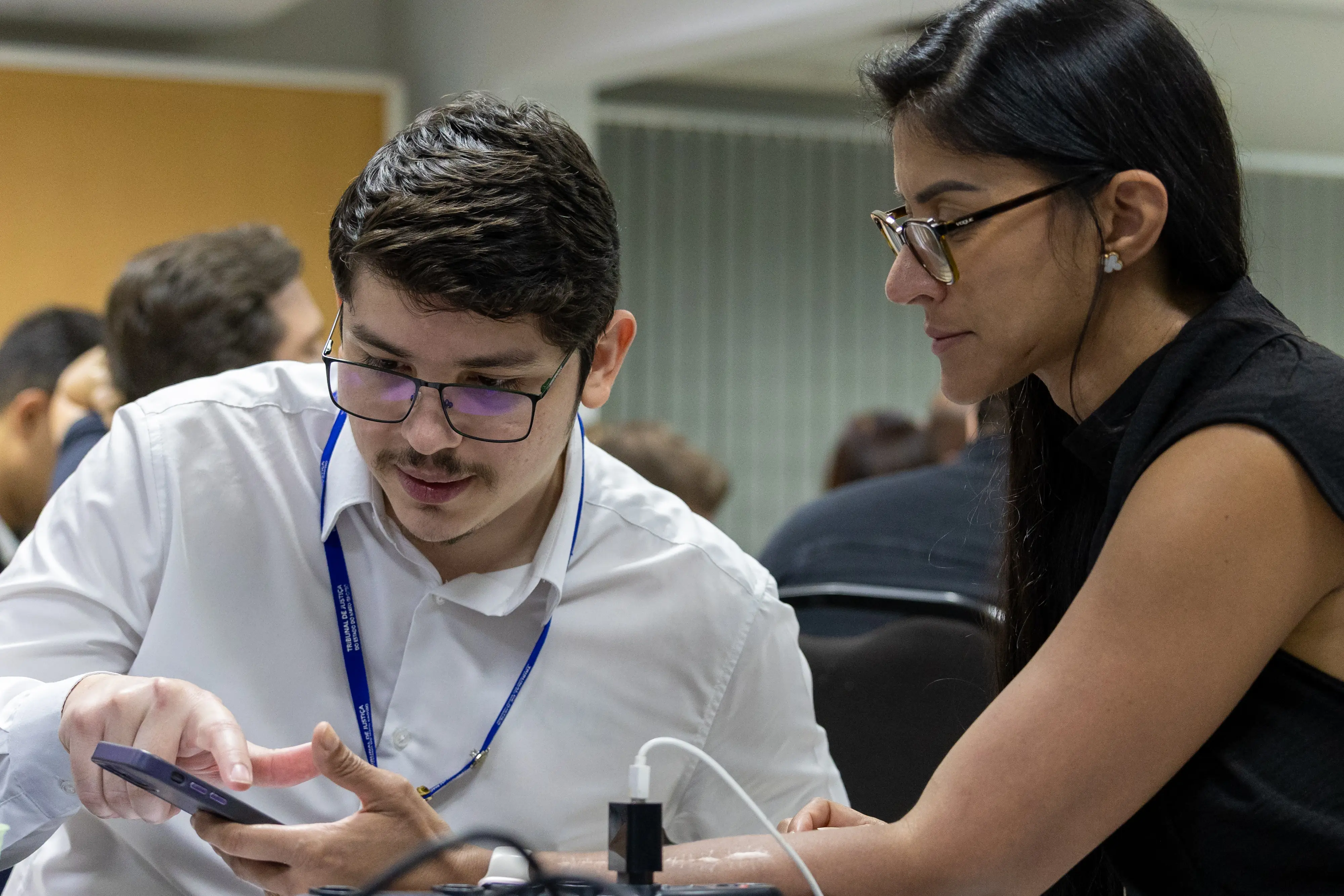 Homem e mulher observam atentamente a tela de um celular. Ele aponta para o aparelho enquanto ela acompanha a explicação, demonstrando cooperação na resolução da tarefa.